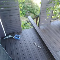 Montaż kompozytowej deski tarasowej w Wałbrzychu: widok z góry na częściowo ukończony taras z ciemnobrązowych desek, w tle zieleń ogrodu, widoczne narzędzia pracy: wiertarka, pojemnik ze śrubami...