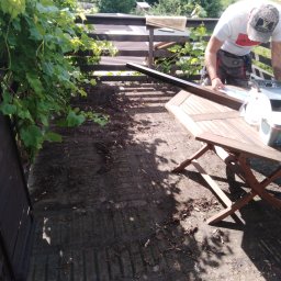 M...K - Pracownik w trakcie montażu drewnianej konstrukcji na zewnątrz, prawdopodobnie elementu zadaszenia, z widoczną drewnianą ramą i narzędziami na składanym stole, w tle zielona roślinność i budynki.