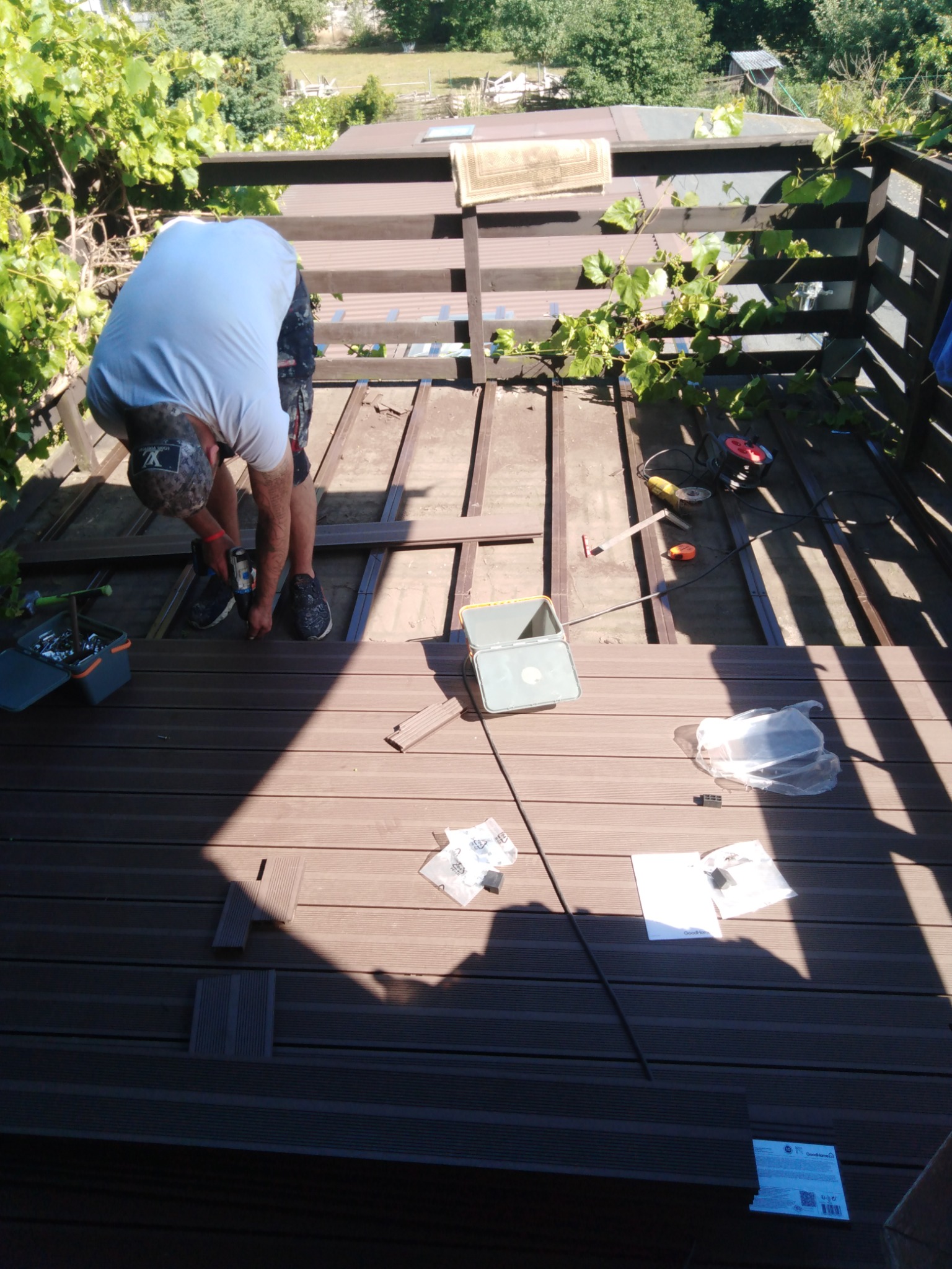 Mężczyzna w czapce i jasnej koszulce montuje deski kompozytowe na tarasie używając wkrętarki, widoczne narzędzia i akcesoria montażowe, w tle roślinność.