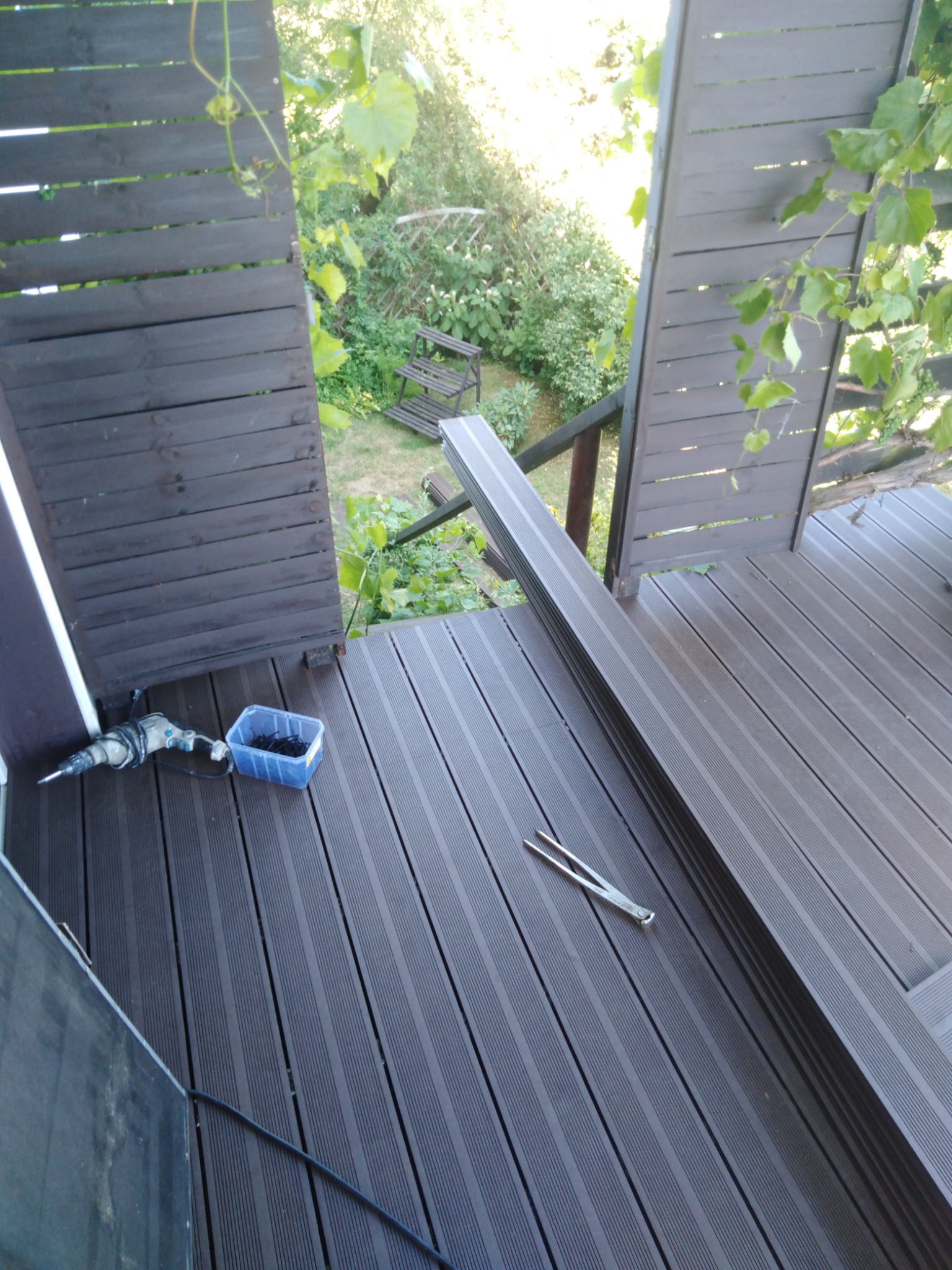 Montaż kompozytowej deski tarasowej w Wałbrzychu: widok z góry na częściowo ukończony taras z ciemnobrązowych desek, w tle zieleń ogrodu, widoczne narzędzia pracy: wiertarka, pojemnik ze śrubami...