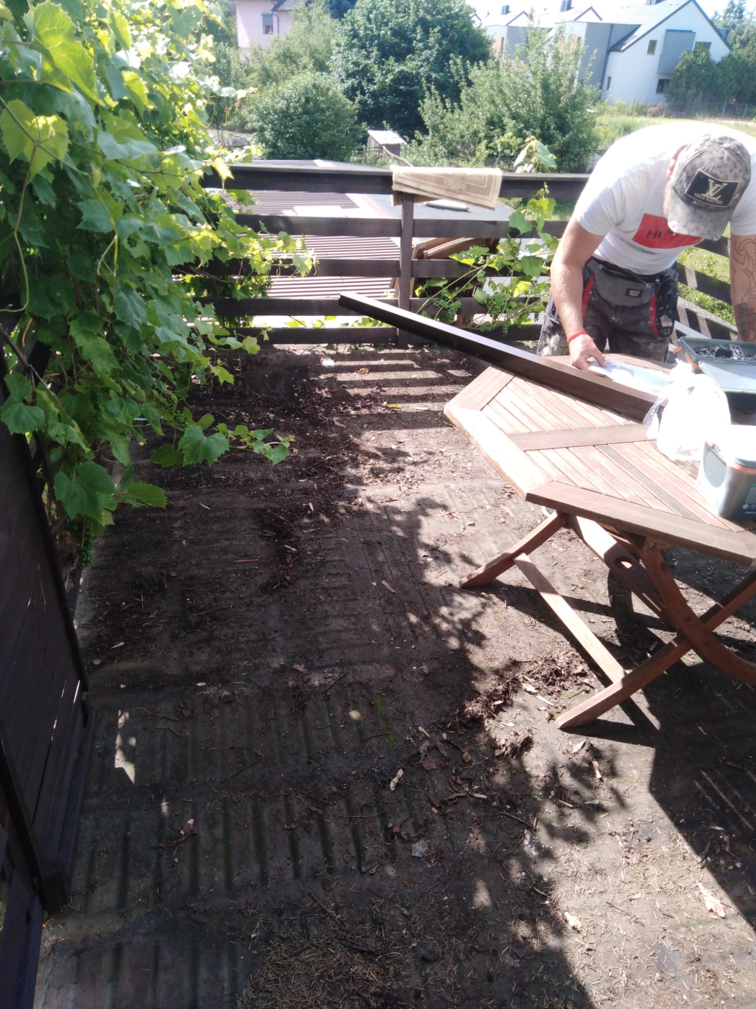 Pracownik w trakcie montażu drewnianej konstrukcji na zewnątrz, prawdopodobnie elementu zadaszenia, z widoczną drewnianą ramą i narzędziami na składanym stole, w tle zielona roślinność i budynki.