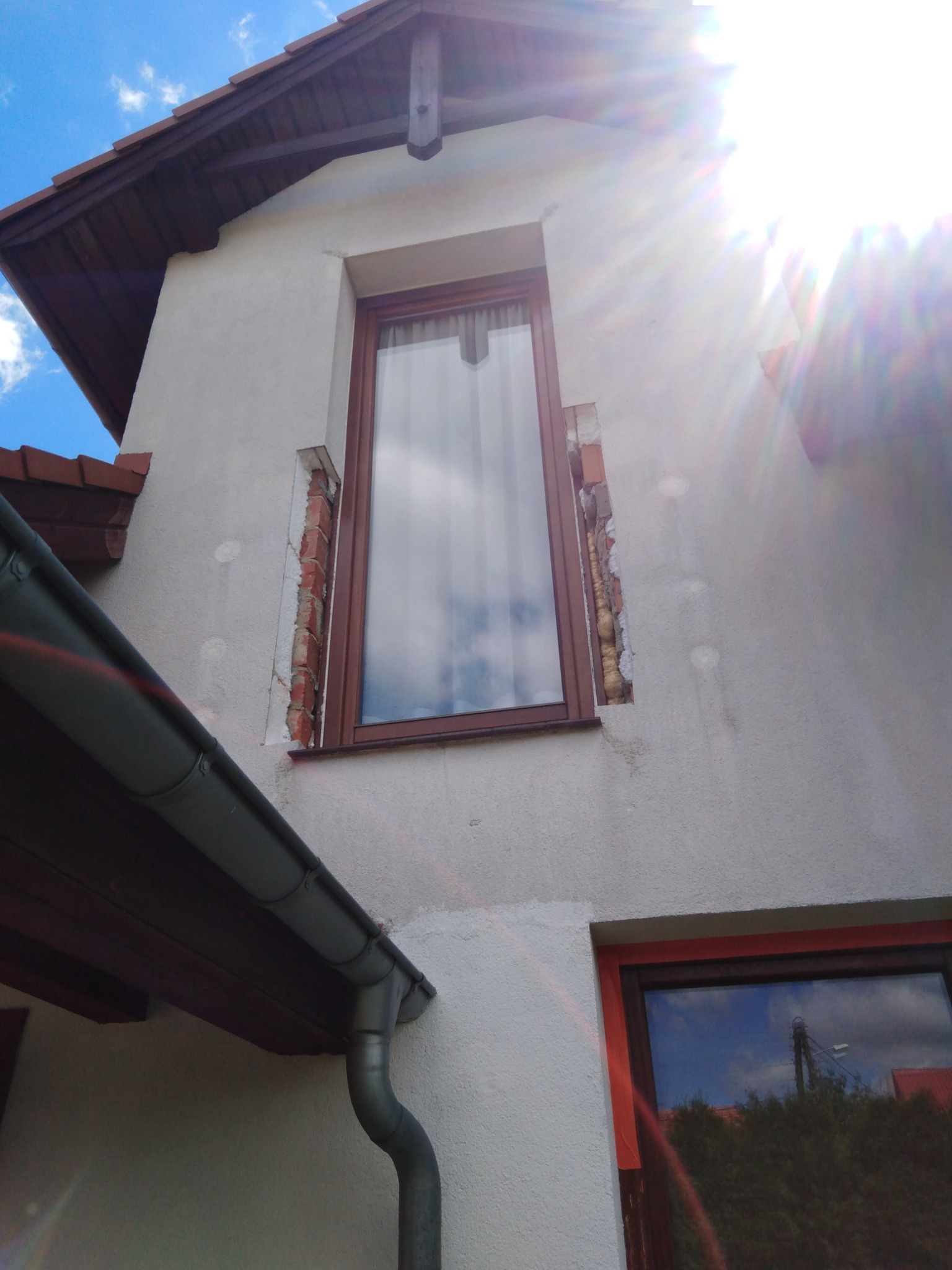 Fragment elewacji budynku z nowo wstawionym oknem; widoczne ślady po usunięciu starej stolarki, odkryta cegła i pianka montażowa, rynna spustowa, jasne tło nieba.