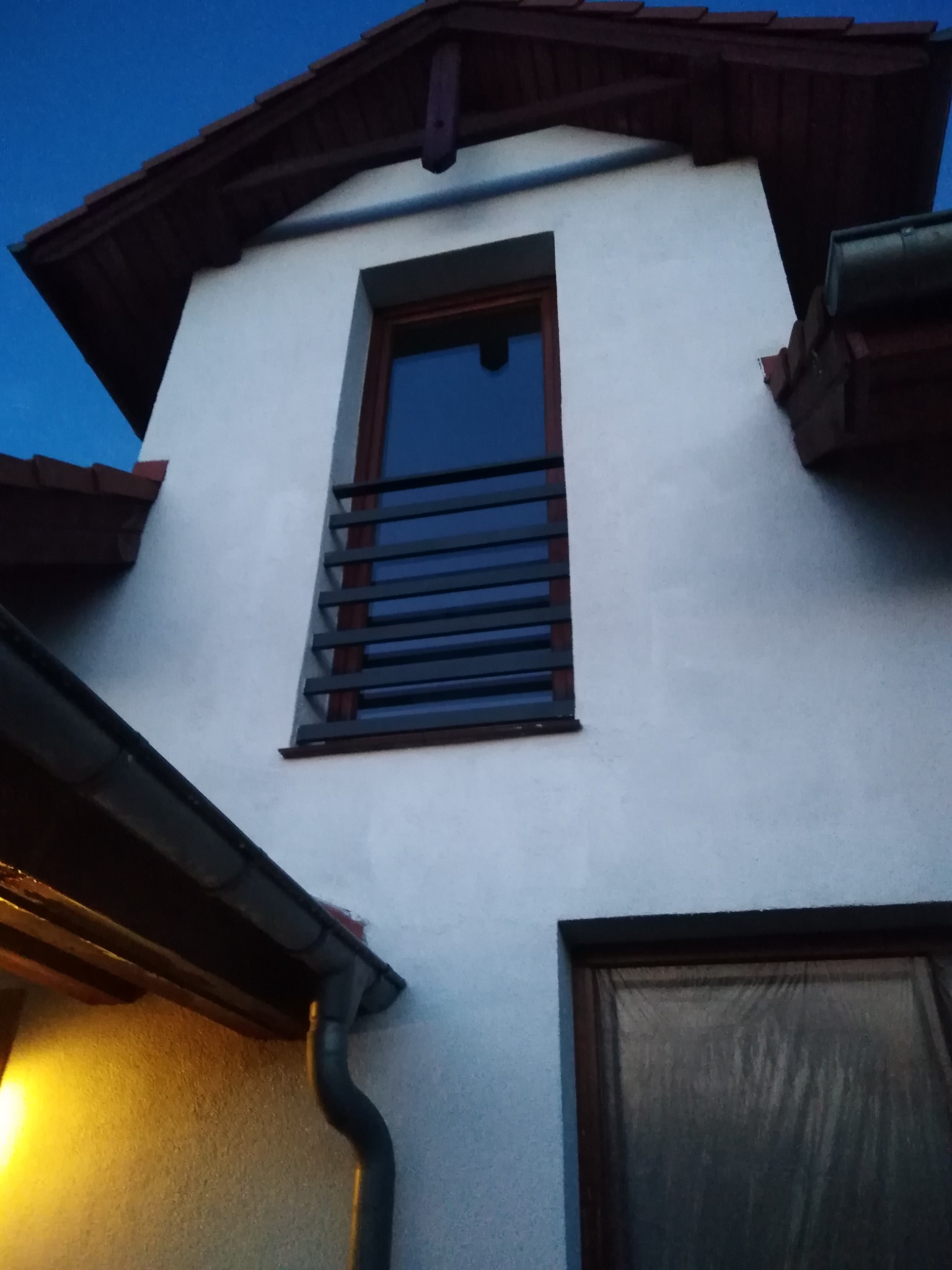 Fragment elewacji domu z oknem z poziomymi szprosami, widoczny fragment dachu z brązową dachówką i rynnami, ujęcie z dołu w pochmurny dzień.