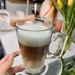 Szklanka latte na spodku, w tle dłoń z delikatnym manicure, lampa UV do paznokci, bukiet tulipanów i kosmetyczka podczas pracy.