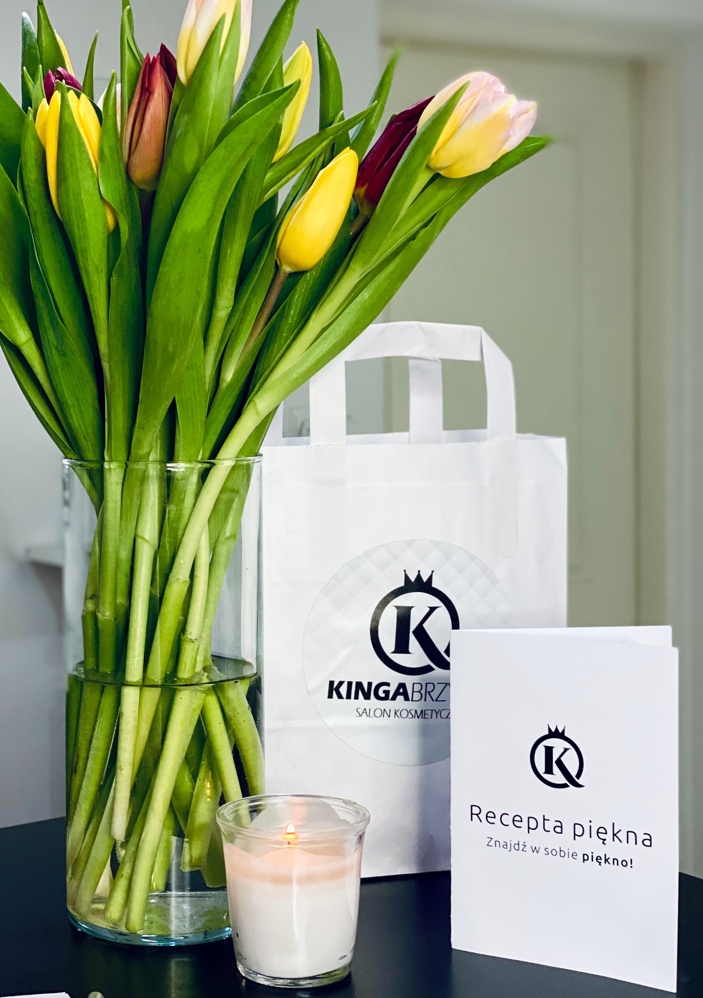 Kompozycja kwiatowa z tulipanami w szklanym wazonie, zapalona świeczka w szklanym pojemniku, torba prezentowa i ulotka z logo salonu kosmetycznego na ciemnym blacie.