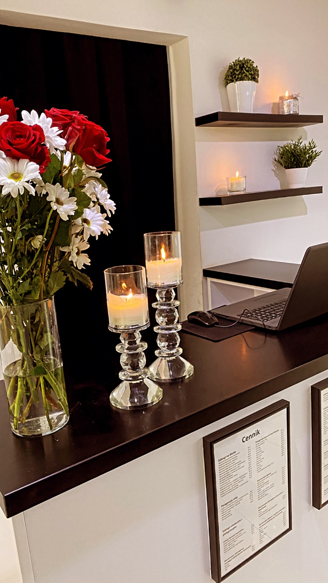 Dekoracyjny stół w salonie kosmetycznym z bukietem czerwonych róż i białych margaretek w szklanym wazonie, dwoma zapalonymi świecami w szklanych świecznikach, laptopem oraz cennikami usług...