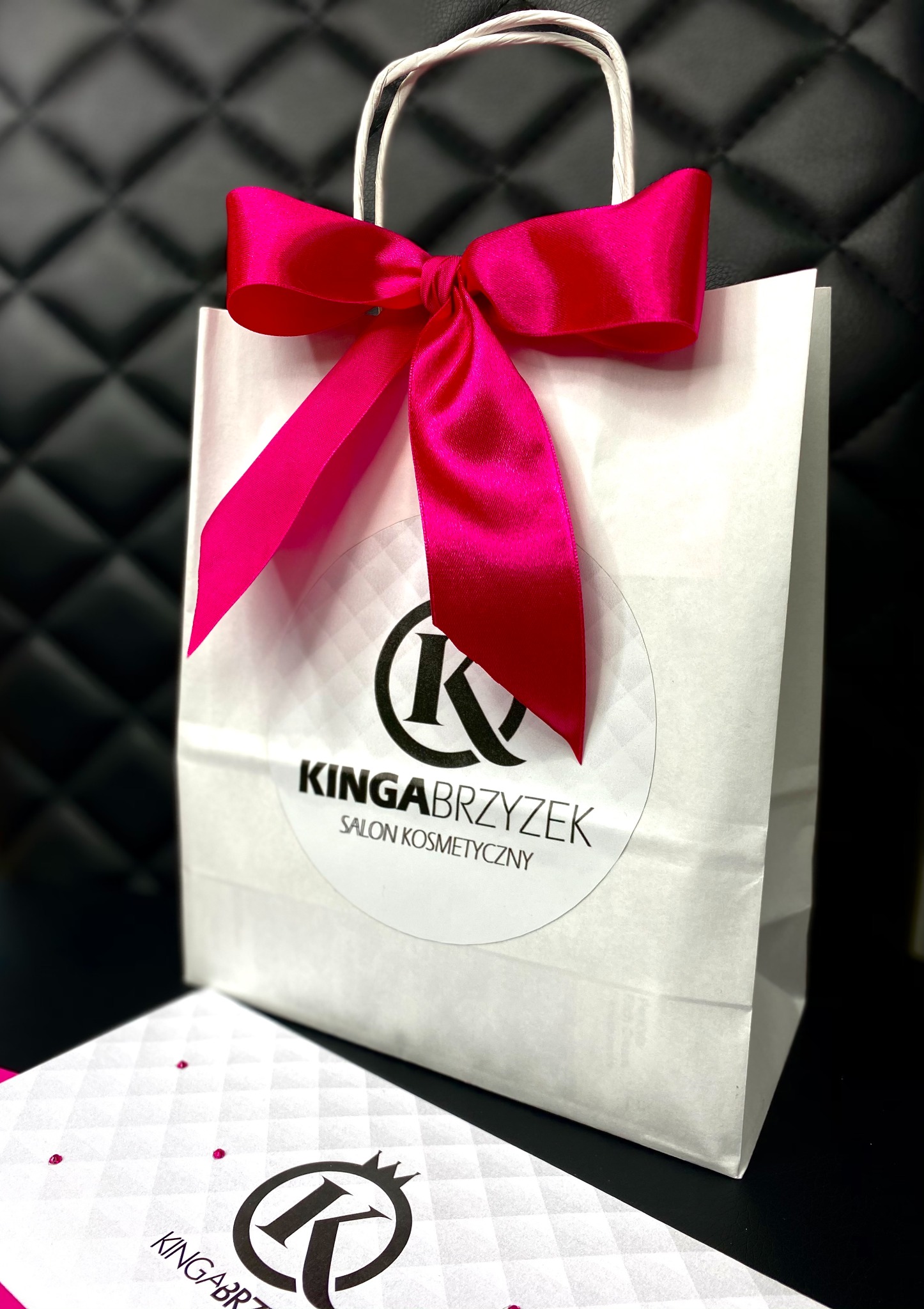 Biała torba prezentowa z różową kokardą i logo salonu kosmetycznego Kinga Brzyzek na tle pikowanego czarnego krzesła, obok wizytówka z logo.