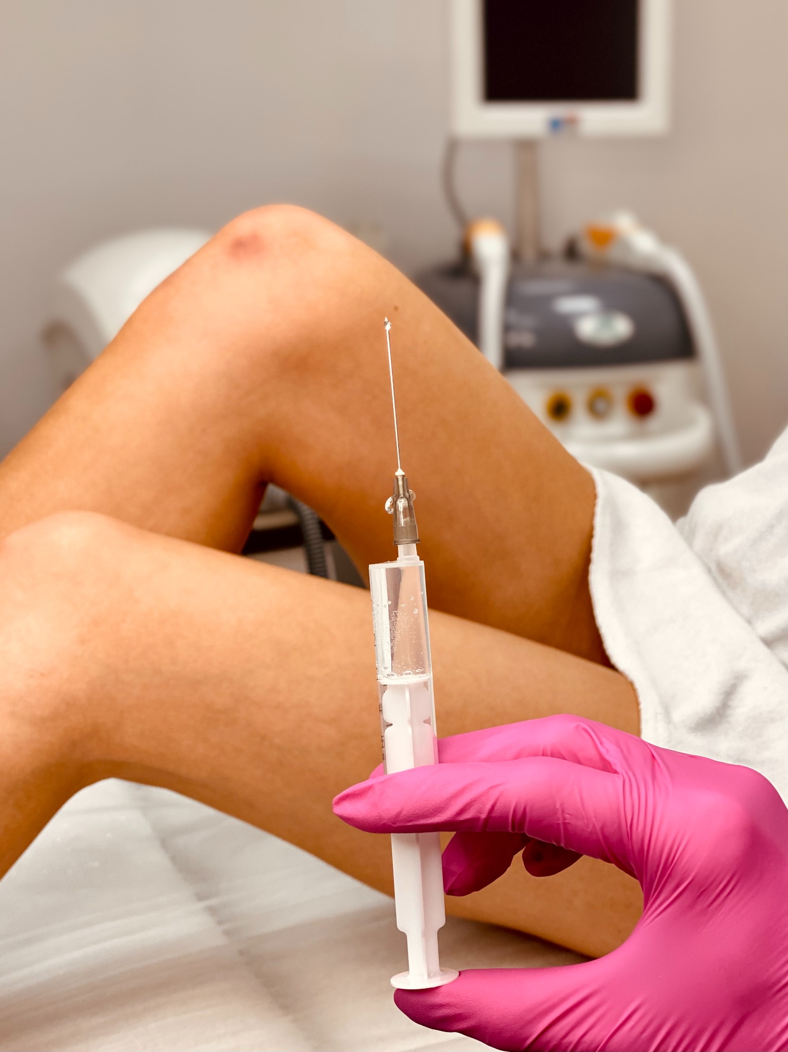 Przygotowanie do iniekcji w okolicy kolana, widoczna igła z kroplą, różowa rękawica, w tle urządzenie do zabiegów.