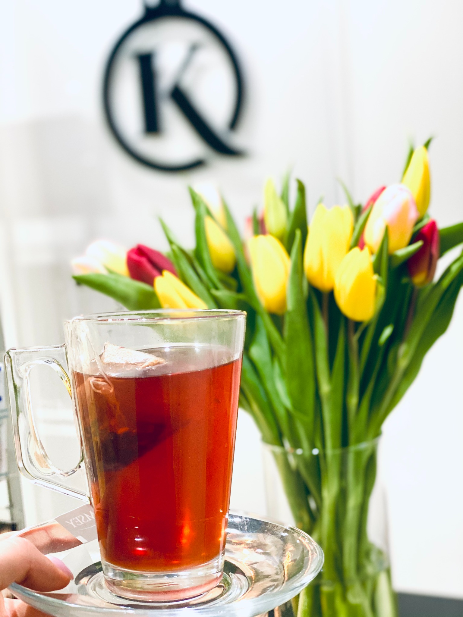 Szklanka herbaty na spodku, trzymana w dłoni, z bukietem tulipanów w tle i logo firmy na ścianie.