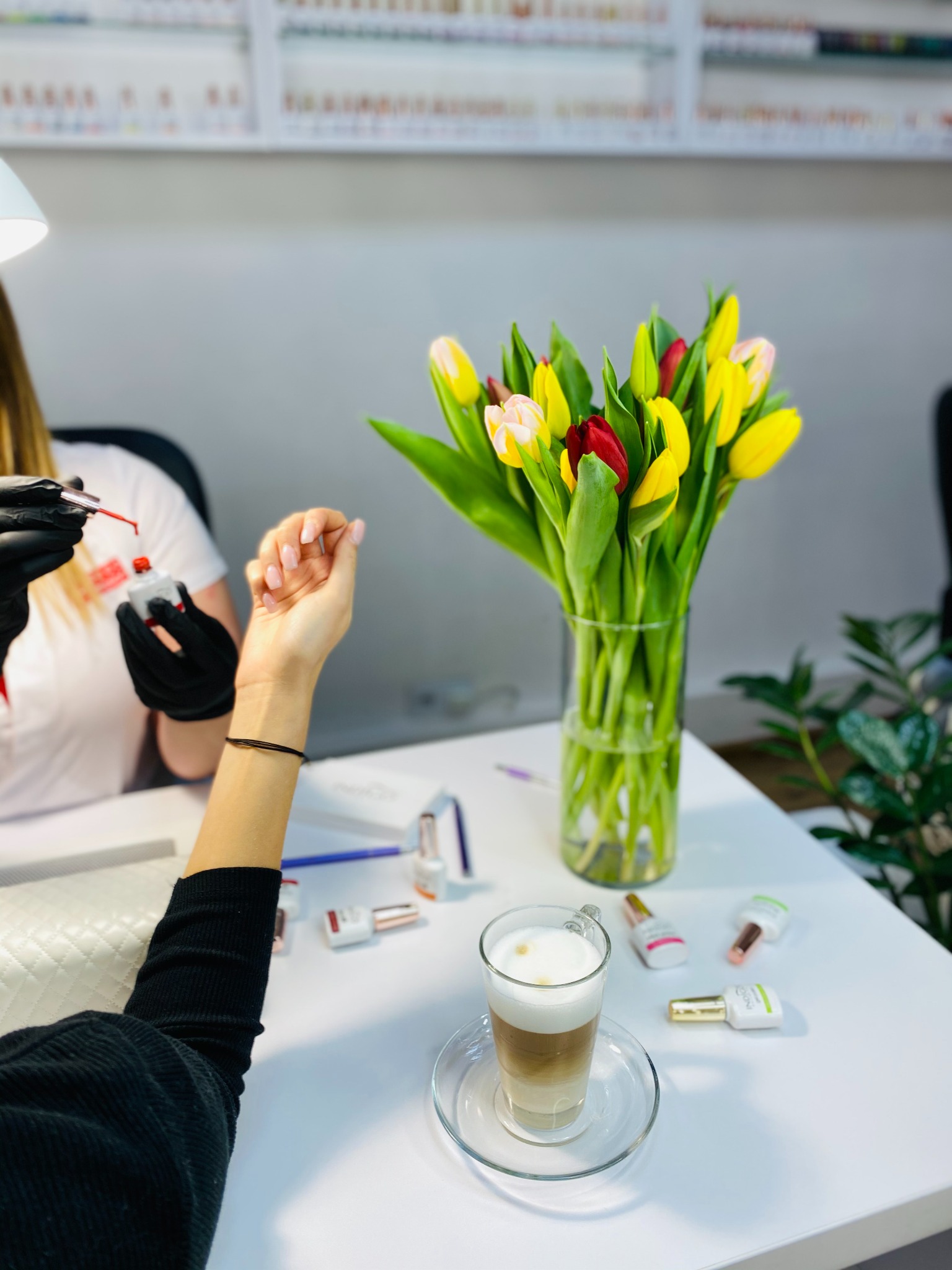 Stylizacja paznokci w salonie: stylistka w czarnych rękawiczkach aplikuje czerwony lakier na paznokcie klientki. Na stole wazon z tulipanami i latte macchiato.