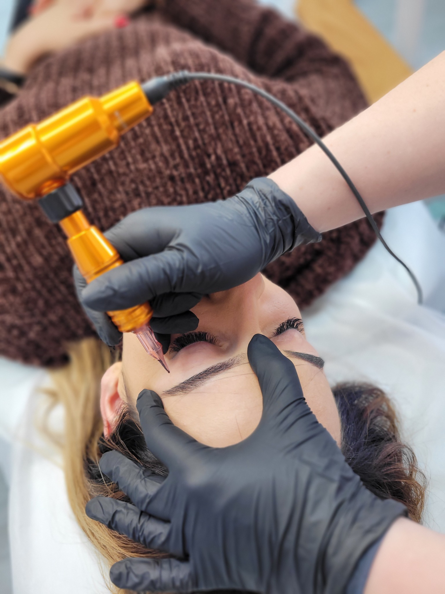 Zabieg pigmentacji brwi wykonywany przez kosmetyczkę w czarnych rękawiczkach przy użyciu złotej maszynki do makijażu permanentnego.