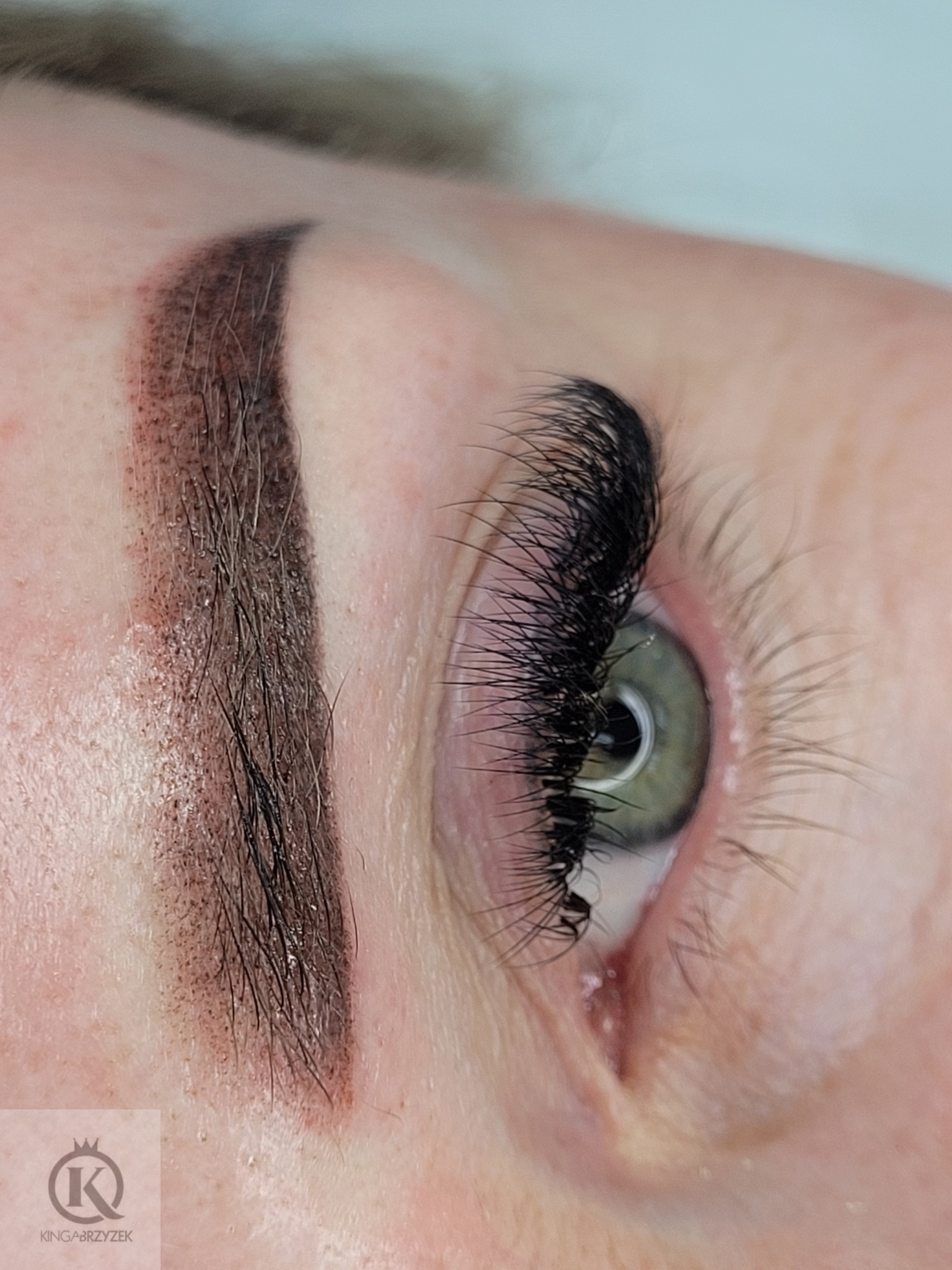 Zbliżenie na oko z zieloną tęczówką, z gęstymi, czarnymi przedłużonymi rzęsami i świeżo wykonanym makijażem permanentnym brwi metodą cieniowania.