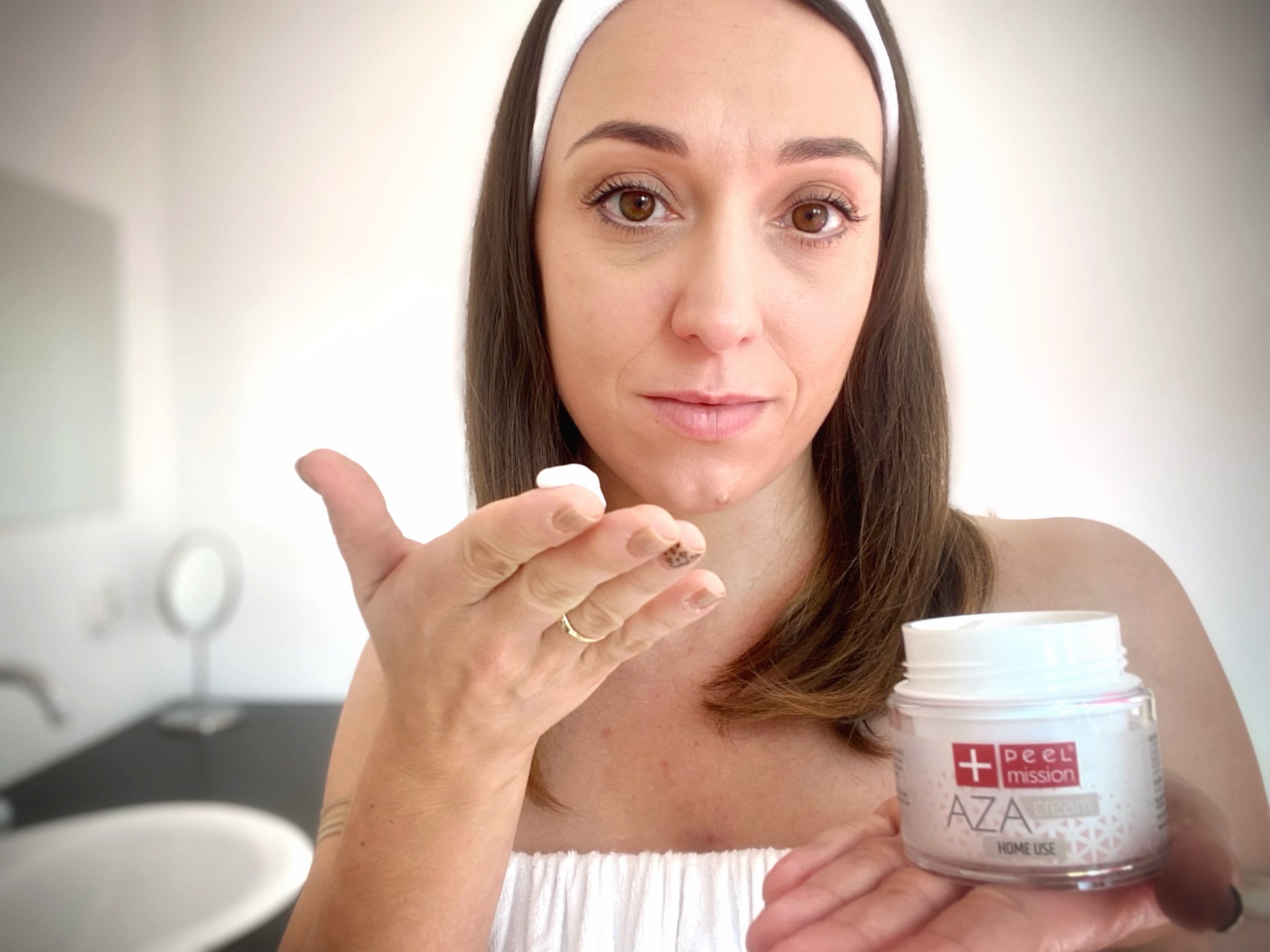 Kobieta z opaską na włosach prezentuje krem AZA Peel Mission trzymany w dłoni, na palcu wskazującym ma nałożoną porcję kremu, w tle widoczna umywalka w łazience.