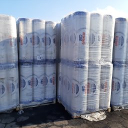 P.W. MBM Marek Moderacki - Palety z rolkami wełny szklanej URSA Amber 33, owinięte folią, składowane na zewnątrz pod błękitnym niebem.