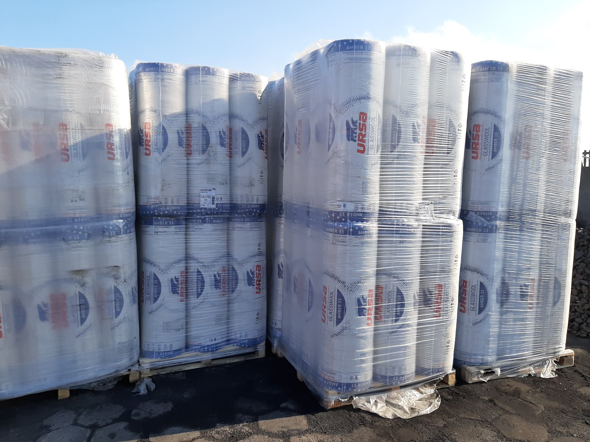 Palety z rolkami wełny szklanej URSA Amber 33, owinięte folią, składowane na zewnątrz pod błękitnym niebem.