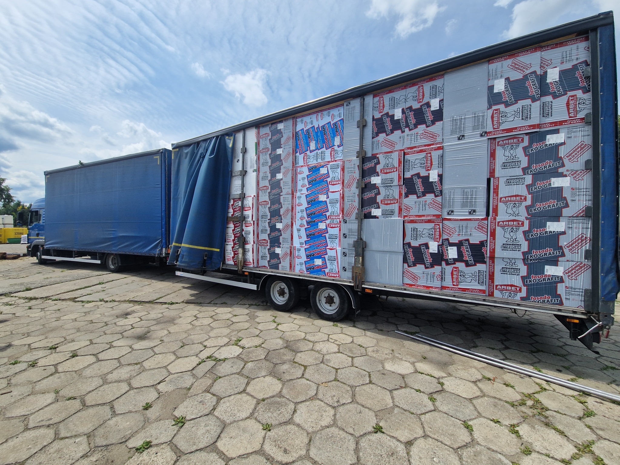 Ciężarówka z naczepą załadowana paczkami materiałów ociepleniowych, marki Arbet, na placu wyłożonym betonową kostką. Widoczne niebieskie plandeki i błękitne niebo z chmurami.