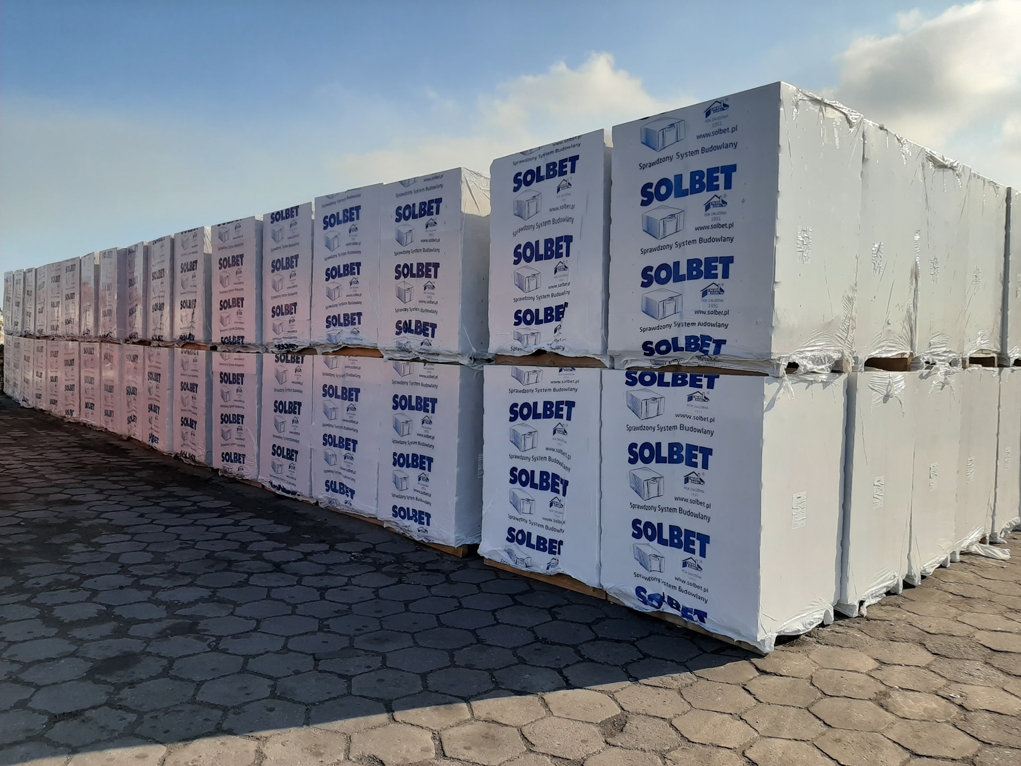 Palety z białymi paczkami bloczków Solbet na placu budowlanym, widoczne niebo i kostka brukowa.