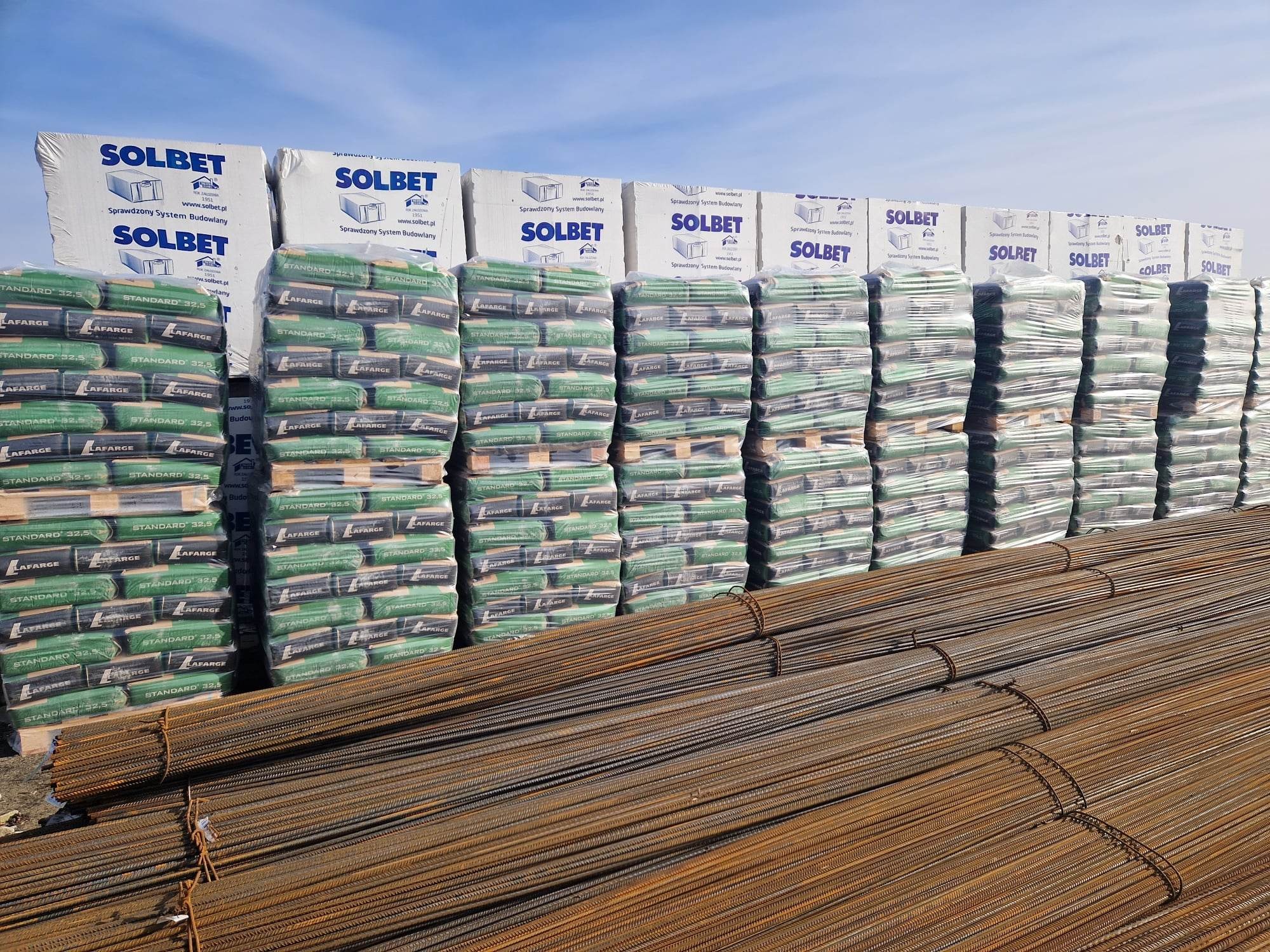 Skladowane materiały budowlane: palety z workami cementu Lafarge Standard 32,5 oraz paczki bloczków Solbet na tle wiązek prętów zbrojeniowych pokrytych rdzą.