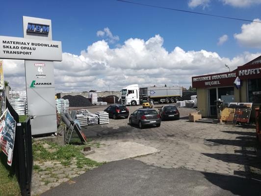 Skład budowlany MBM z zaparkowanymi samochodami, stertami materiałów budowlanych i węgla, oraz ciężarówką w tle pod błękitnym niebem z chmurami.