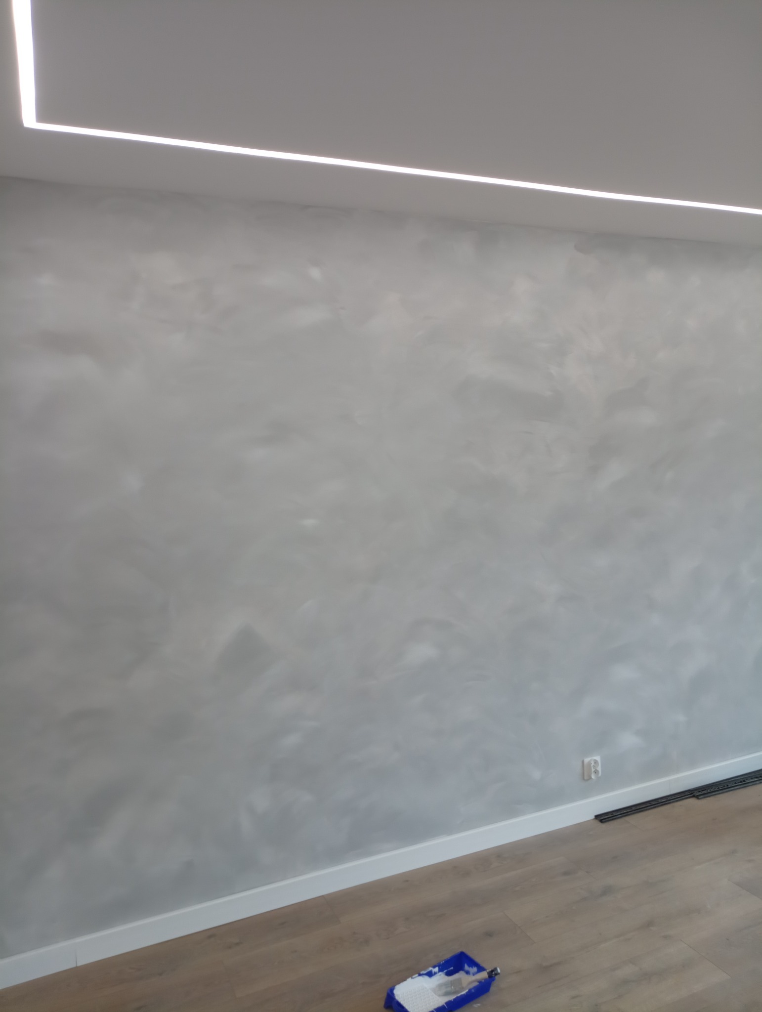 Ściana z efektem dekoracyjnym w odcieniach szarości, oświetlona ledowym paskiem na suficie, widoczna puszka z farbą i wałkiem na podłodze w Bukowcu.