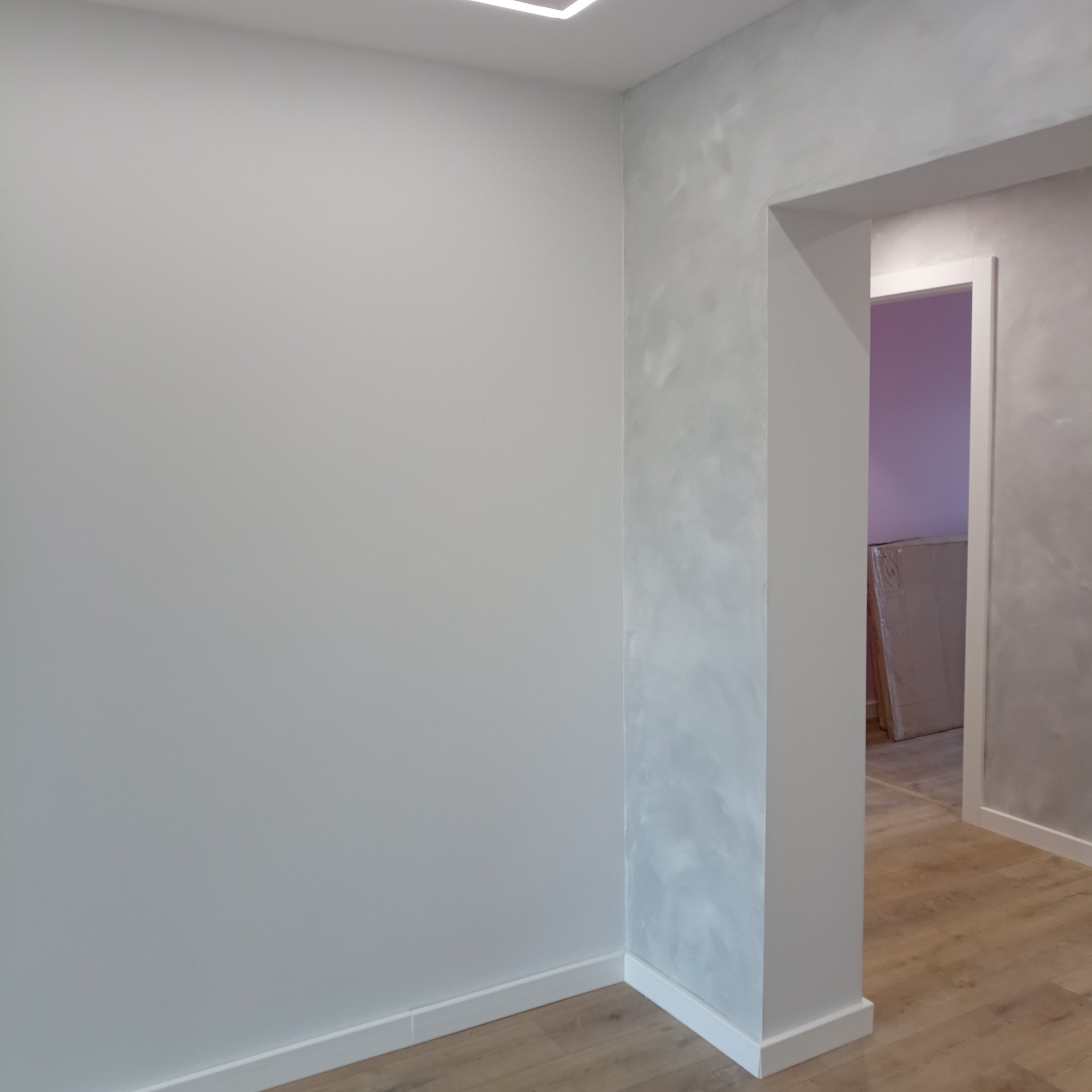 Pomalowane jasnoszare ściany z efektem dekoracyjnym, widoczna podłoga z paneli oraz fragment przejścia do innego pomieszczenia, gdzie oparta jest o ścianę zapakowana płyta.