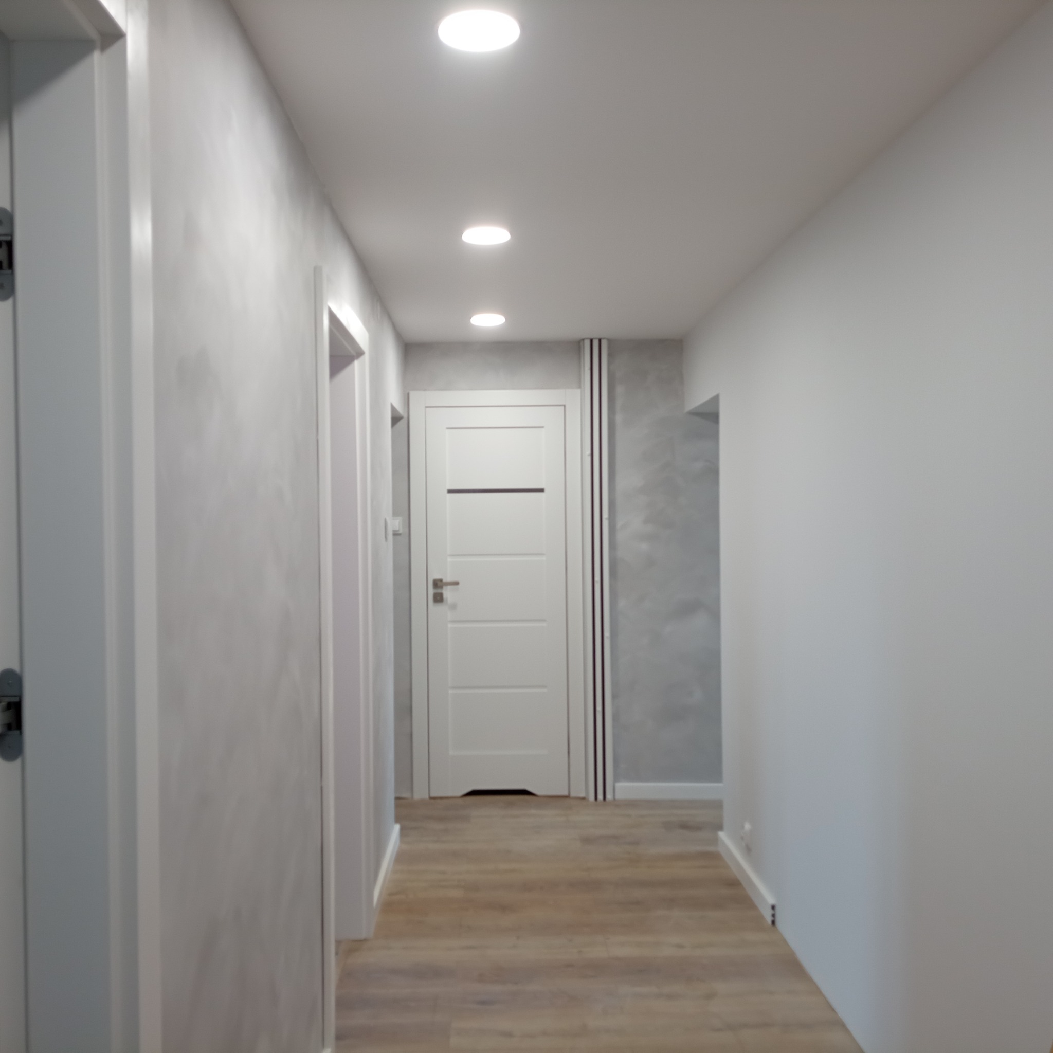 Jasny korytarz po remoncie: drewnopodobne panele podłogowe, białe ściany z szarym akcentem strukturalnym, białe drzwi z poziomymi przeszkleniami, oświetlenie sufitowe punktowe.