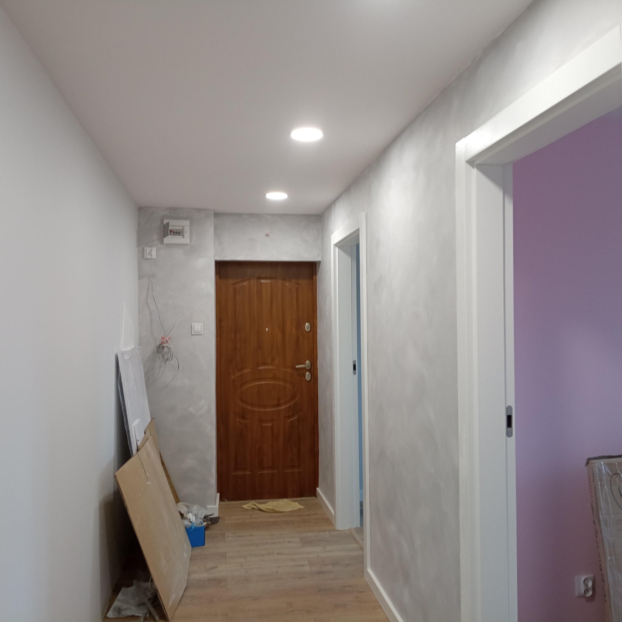 Korytarz po malowaniu ścian na szaro z efektem dekoracyjnym, widoczne drzwi wejściowe i wejście do pokoju pomalowanego na fioletowo, na podłodze panele, przy ścianie narzędzia i materiały budowlane.