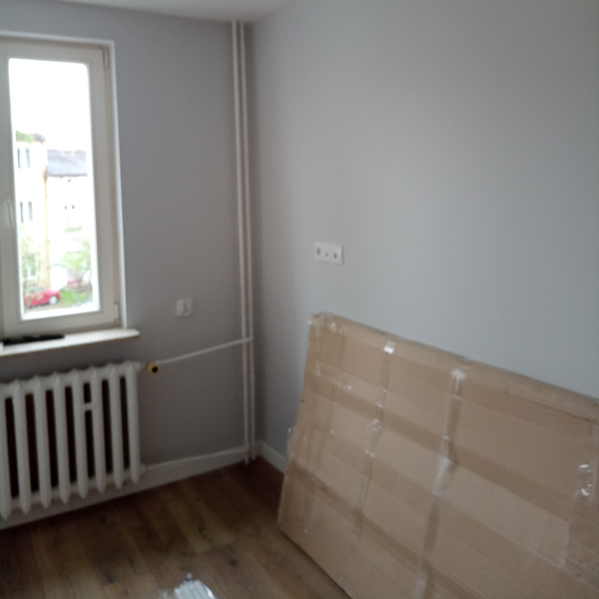 Pokój przed odświeżeniem: szare ściany, drewniana podłoga, białe okno z widokiem na zieleń, grzejnik żeliwny i kartonowe zabezpieczenie pod ścianą.