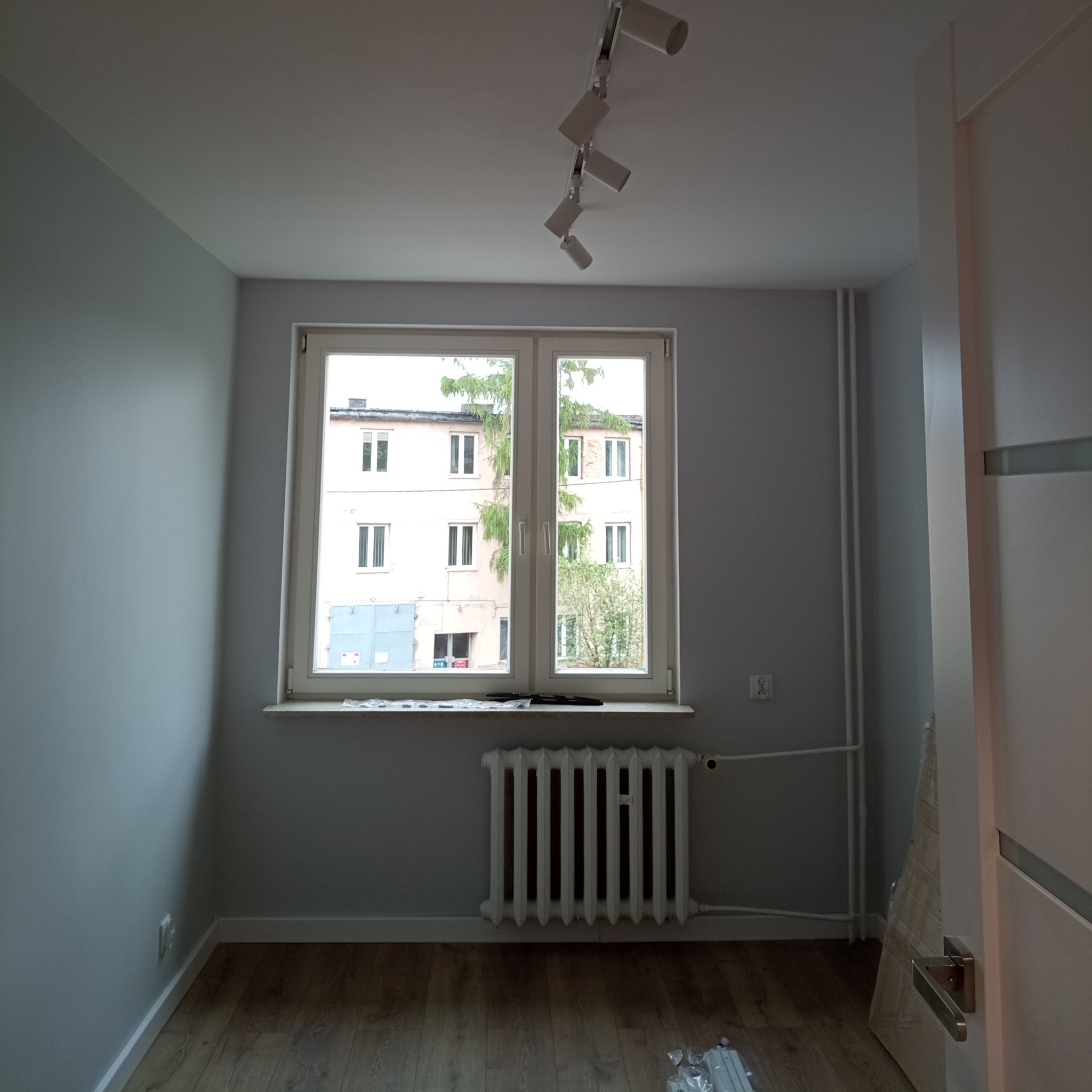 Pomalowane na szaro wnętrze pokoju z drewnianą podłogą, białym grzejnikiem i oknem z widokiem na budynek mieszkalny, oświetlone lampami sufitowymi.