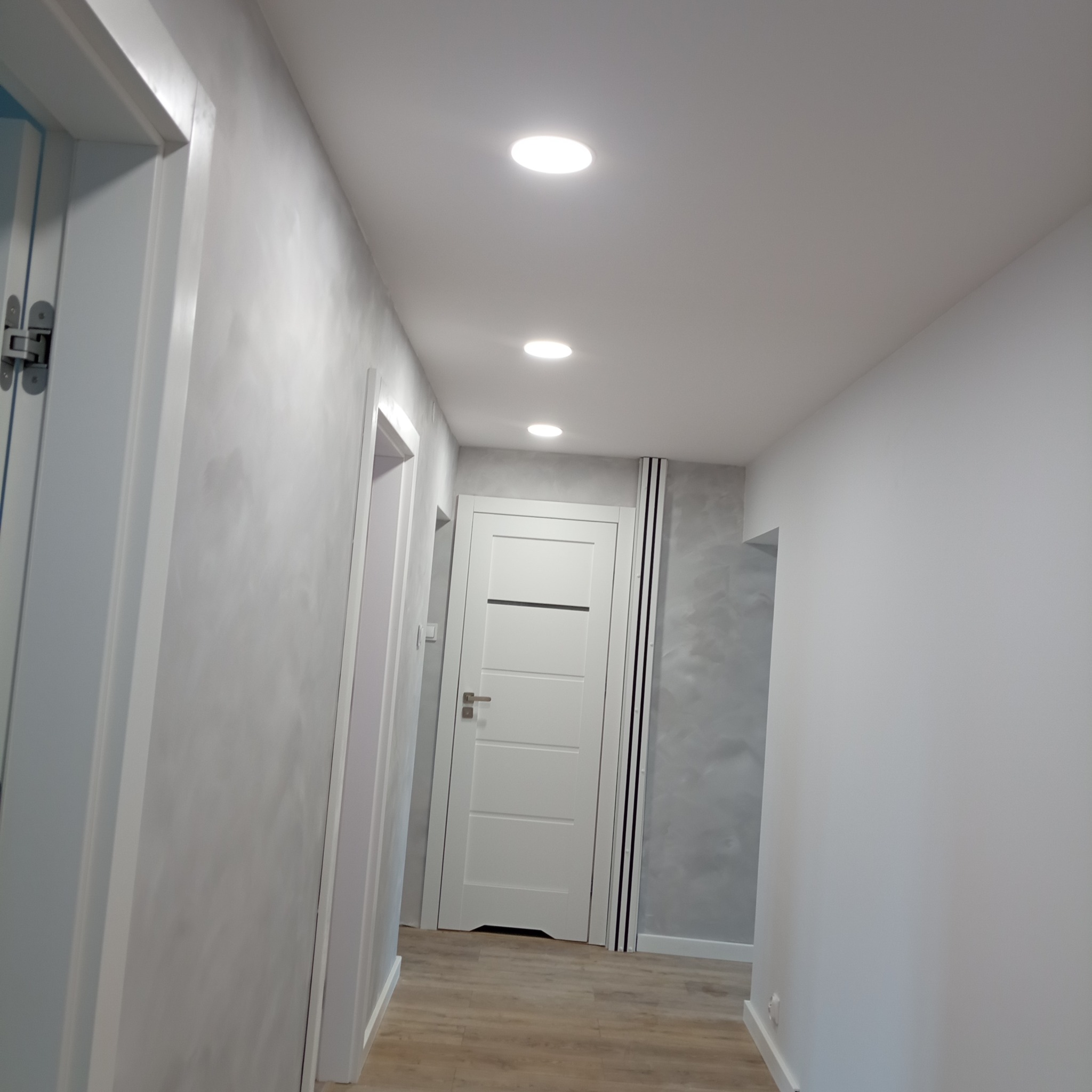 Korytarz po remoncie z jasnymi panelami podłogowymi, ścianami w odcieniach szarości z efektem dekoracyjnym i białymi drzwiami z poziomymi panelami oraz wpuszczanymi lampami sufitowymi.