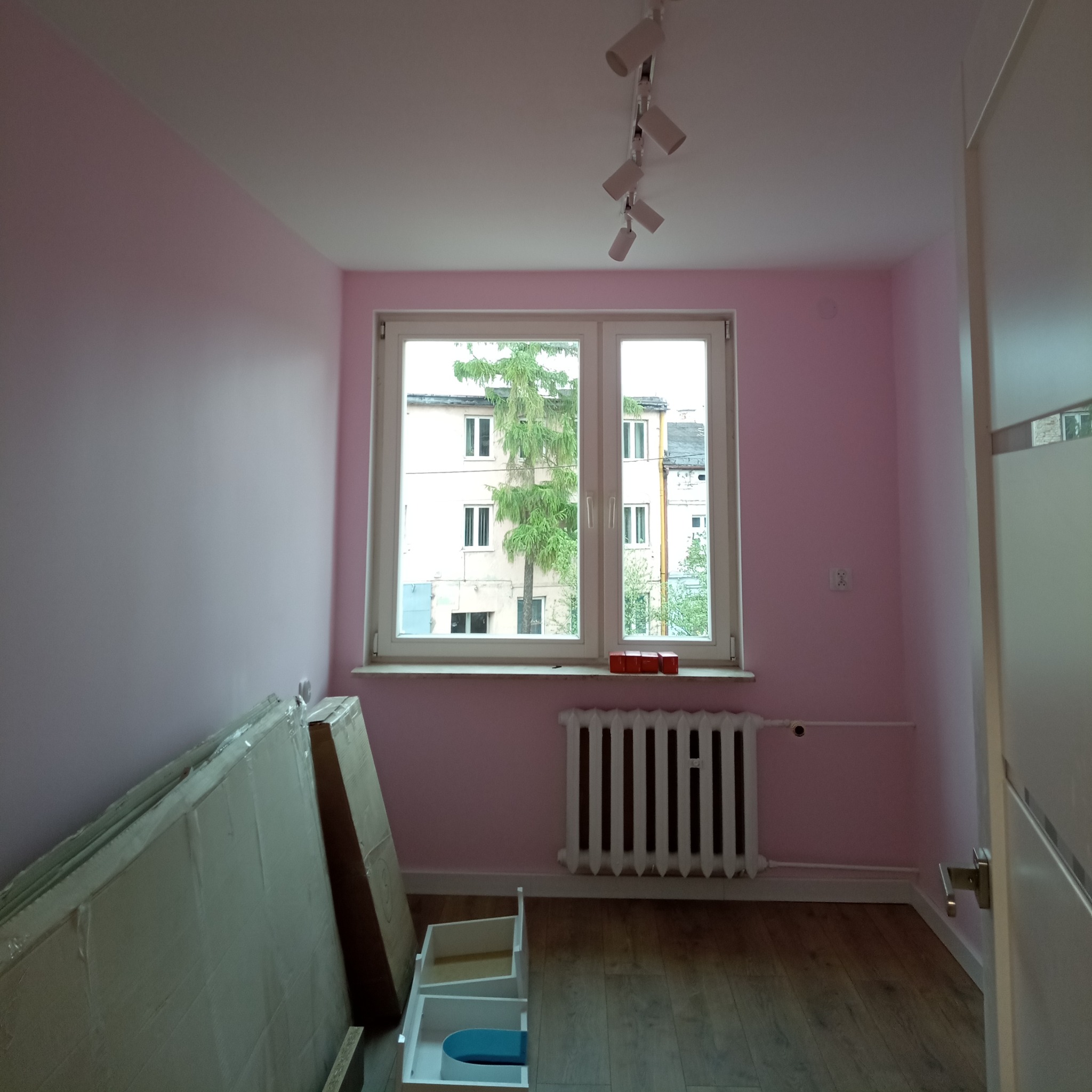 Pomalowany na różowo pokój z drewnianą podłogą, grzejnikiem i oknem z widokiem na drzewo, częściowo umeblowany, z rozpakowanymi elementami mebli przy ścianie.