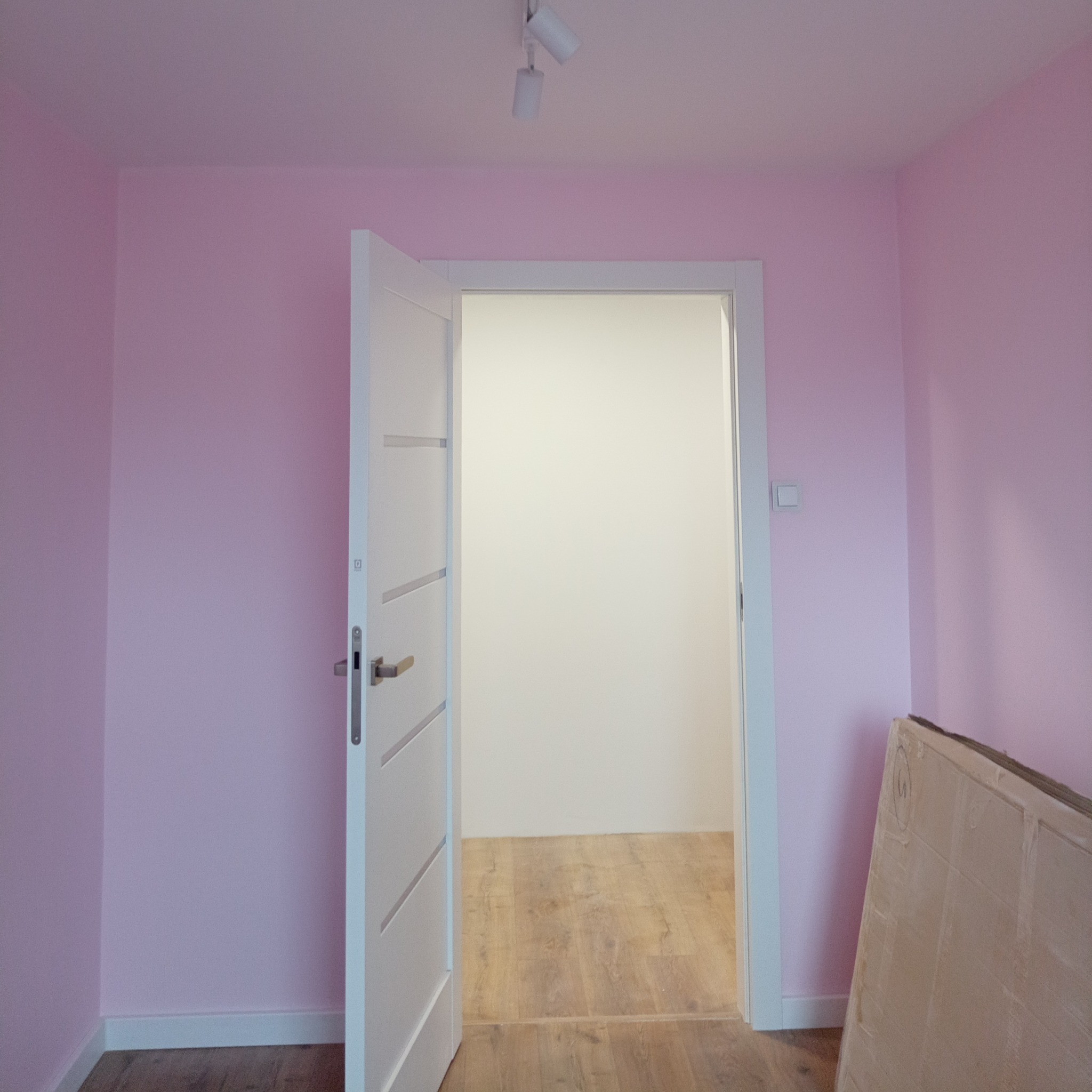 Różowe ściany świeżo pomalowanego pokoju z otwartymi białymi drzwiami i panelami na podłodze, widoczne panele podłogowe ocieplone folią, z sufitu zwisają dwa reflektory.