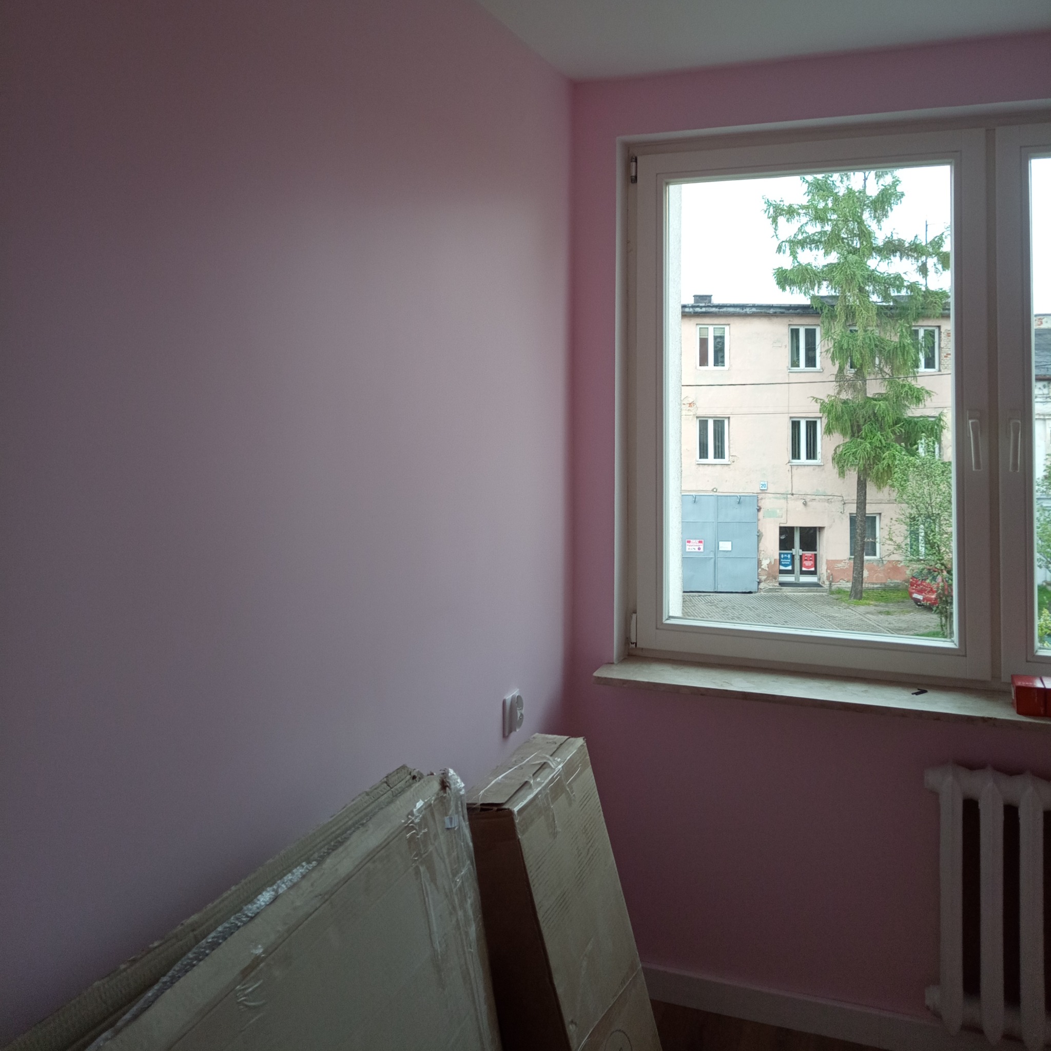 Świeżo pomalowane na różowo wnętrze pokoju z oknem wychodzącym na podwórko z budynkiem i drzewem; w rogu stoją kartony z panelami podłogowymi.