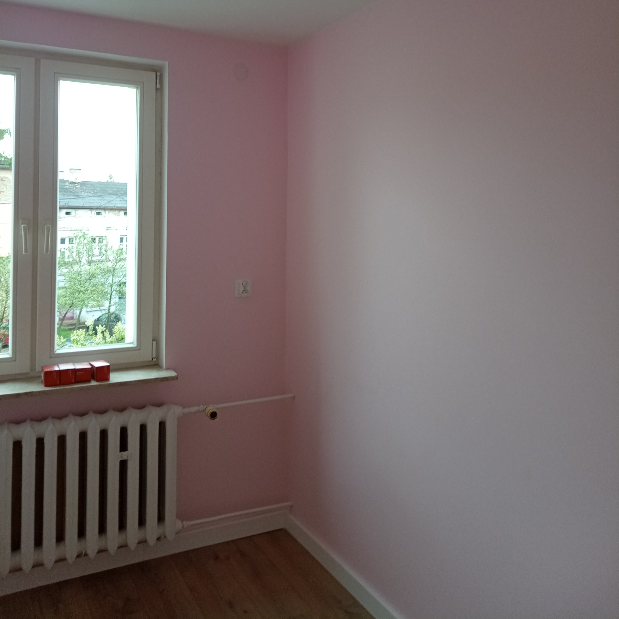 Pokój po malowaniu na różowo, z widocznym białym grzejnikiem, drewnianą podłogą i oknem z widokiem na budynki i drzewa; na parapecie leżą cztery czerwone klocki.