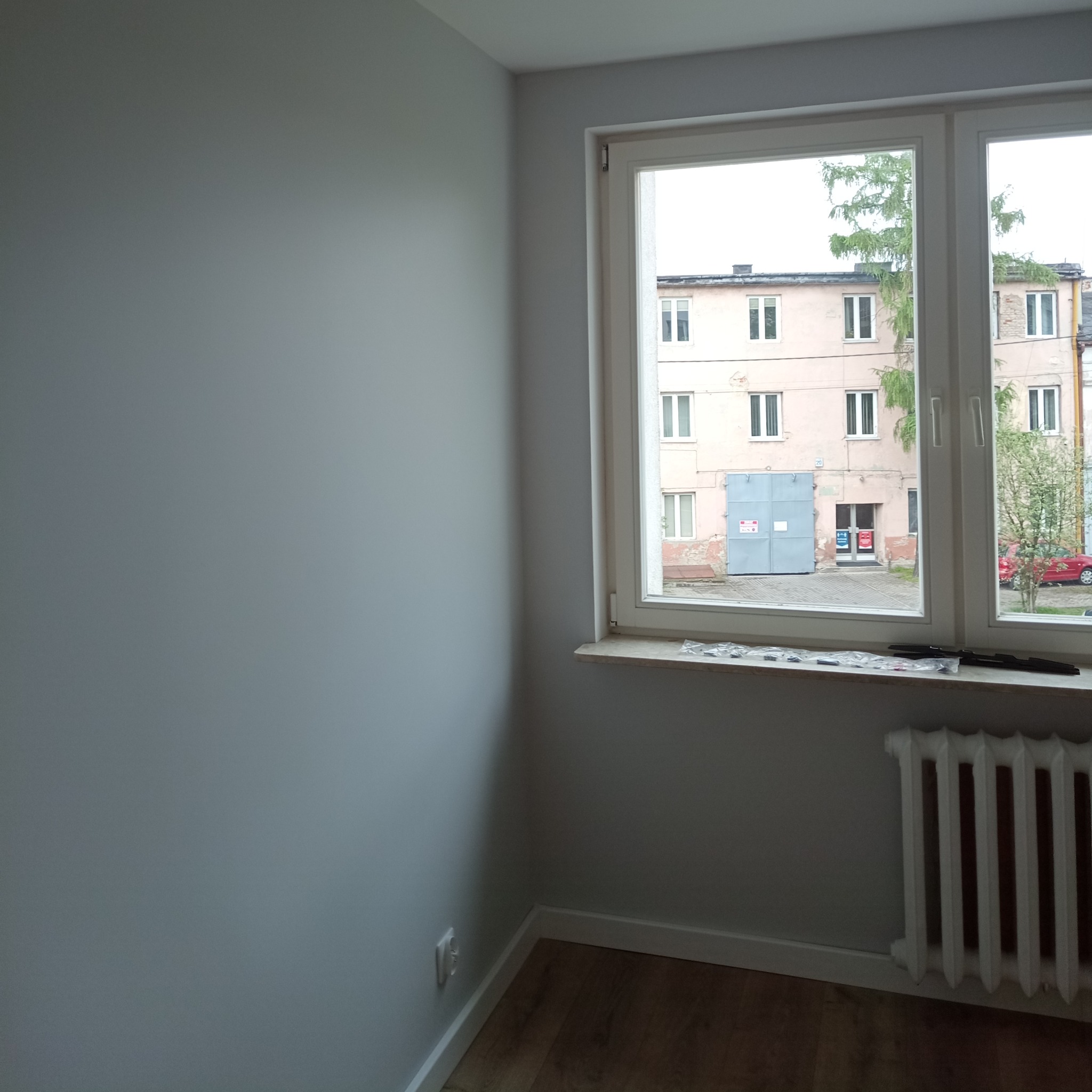 Pomalowane na szaro ściany pokoju z drewnianą podłogą, widok przez okno na budynek mieszkalny i garaż z niebieskimi drzwiami, na parapecie leżą listwy przypodłogowe do montażu, a pod oknem grzejnik.