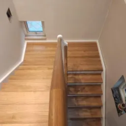 Widok z góry na drewniane schody z jasnego drewna, z prostą, okrągłą poręczą z ciemniejszego drewna i białymi listwami przypodłogowymi, oświetlone naturalnym światłem z okna dachowego.