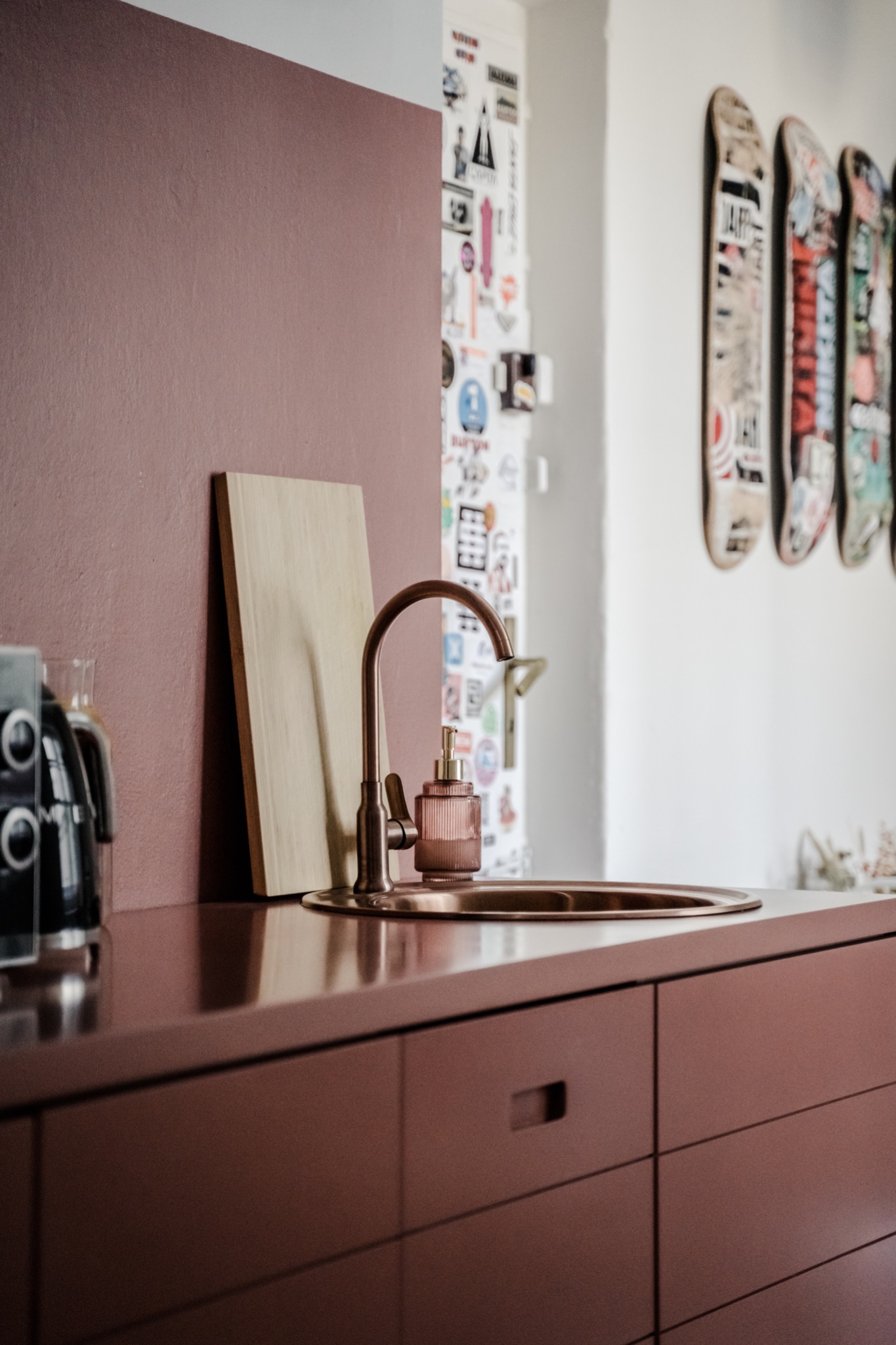 Kuchenny zlew w kolorze terakoty z miedzianą armaturą i dozownikiem mydła w prążkowanym szkle, na tle ściany z naklejkami i deskorolkami.