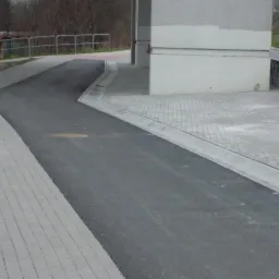 Nowa nawierzchnia z kostki brukowej w szarym odcieniu, połączona z asfaltową drogą, obok betonowego budynku z barierkami.