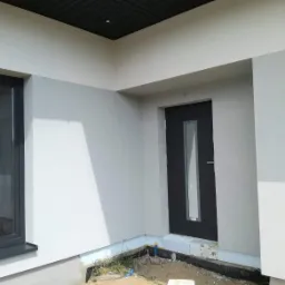 Fragment elewacji budynku po nałożeniu tynku w odcieniach szarości, widoczne okno z ciemną ramą i ciemne drzwi wejściowe z przeszkleniem, podbitka dachowa w kolorze antracytowym.