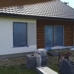 Dom jednorodzinny w trakcie ocieplania elewacji: fragment ściany otynkowanej, okna zabezpieczone folią, obok stos styropianu, wiadro z klejem Kreisel i narzędzia.