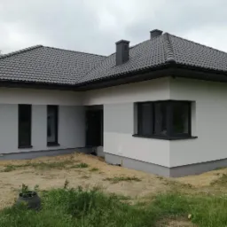 Nowoczesny dom jednorodzinny z szarą elewacją, ciemnymi oknami i grafitową dachówką, częściowo zagospodarowany teren wokół budynku.