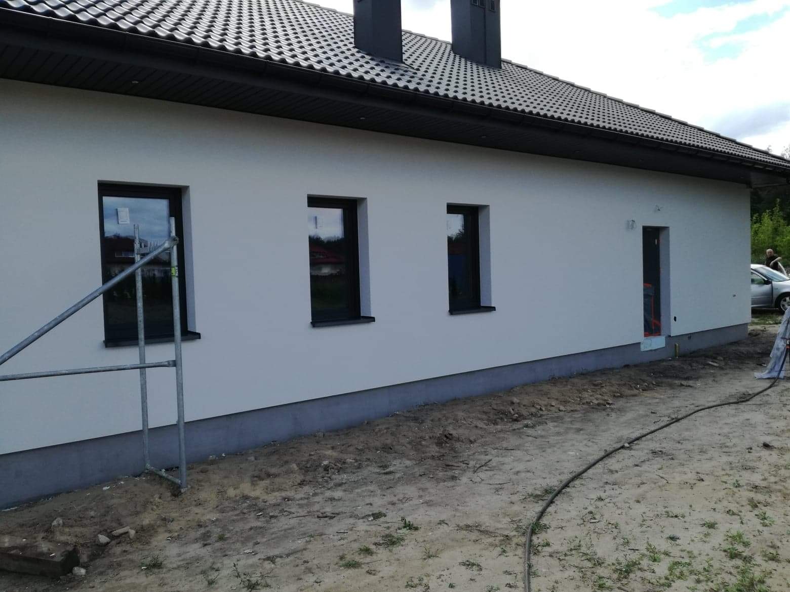 Biała elewacja domu jednorodzinnego z czarnymi oknami i dachem, widoczne rusztowanie przy oknie, szara podmurówka i częściowo zagospodarowany teren wokół budynku.