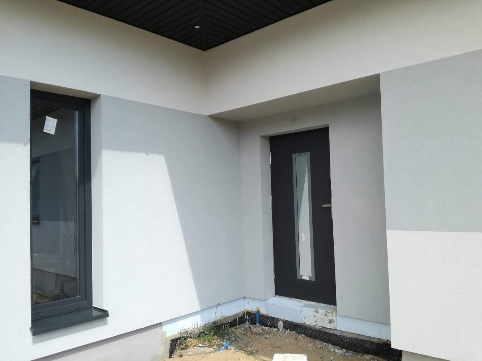 Fragment elewacji budynku po nałożeniu tynku w odcieniach szarości, widoczne okno z ciemną ramą i ciemne drzwi wejściowe z przeszkleniem, podbitka dachowa w kolorze antracytowym.