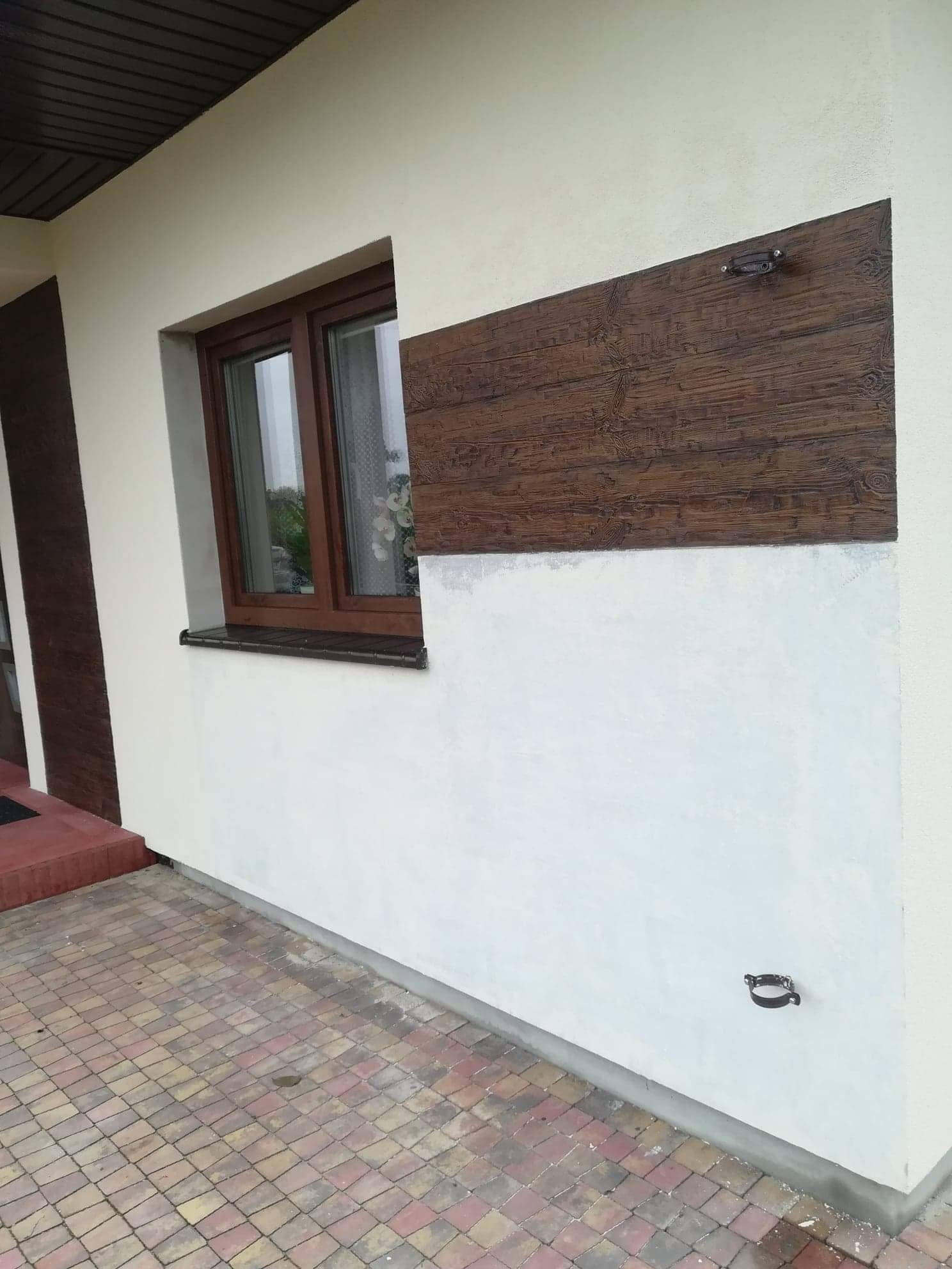Fragment elewacji domu z drewnopodobnym panelem dekoracyjnym, częściowo pomalowany na biało, widoczne okno z brązową ramą i kostka brukowa na podłożu.