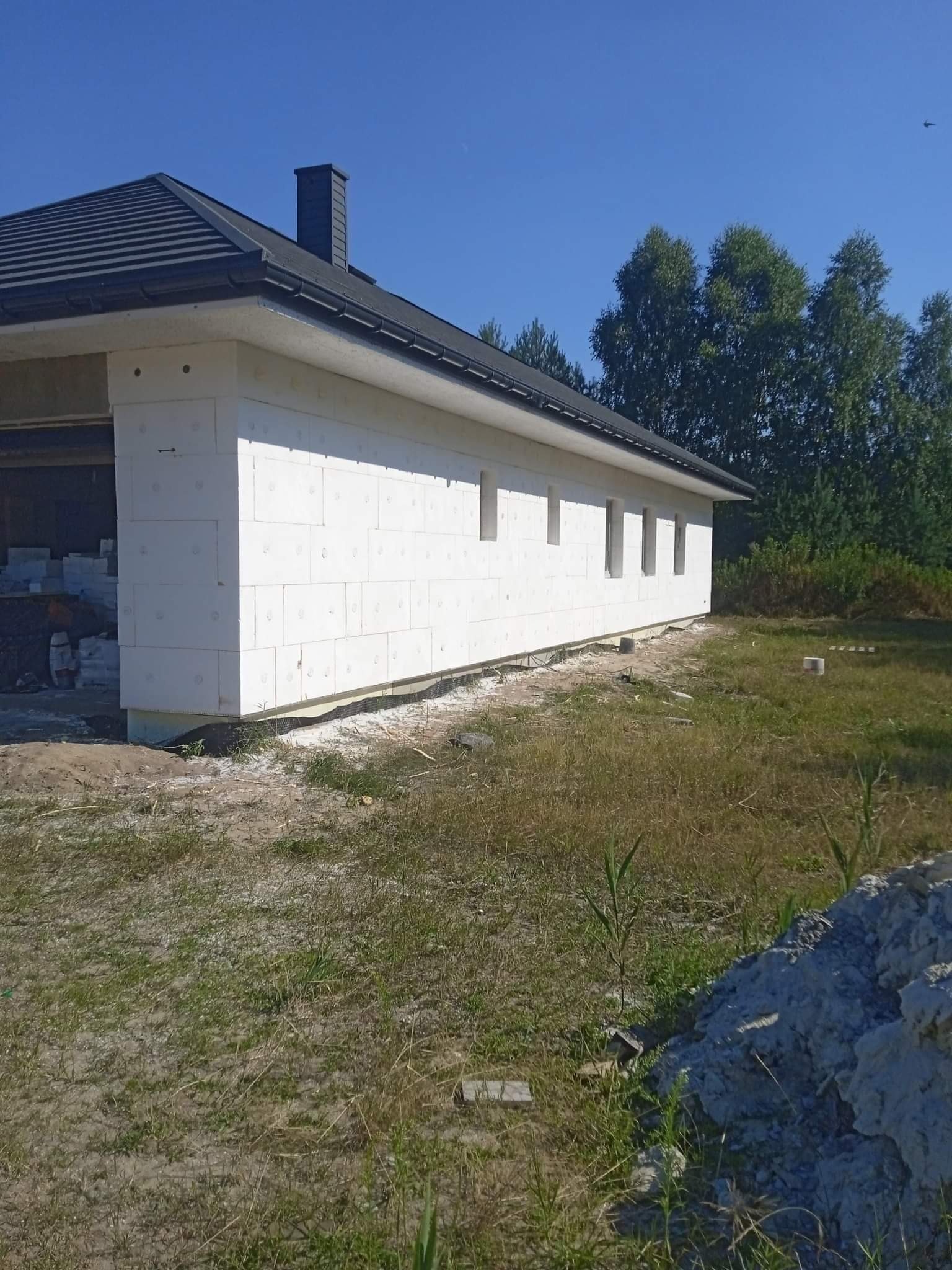 Budynek w trakcie izolacji zewnętrznej, widoczne białe płyty izolacyjne na ścianach, czarny dach i komin, teren wokół w stanie surowym.