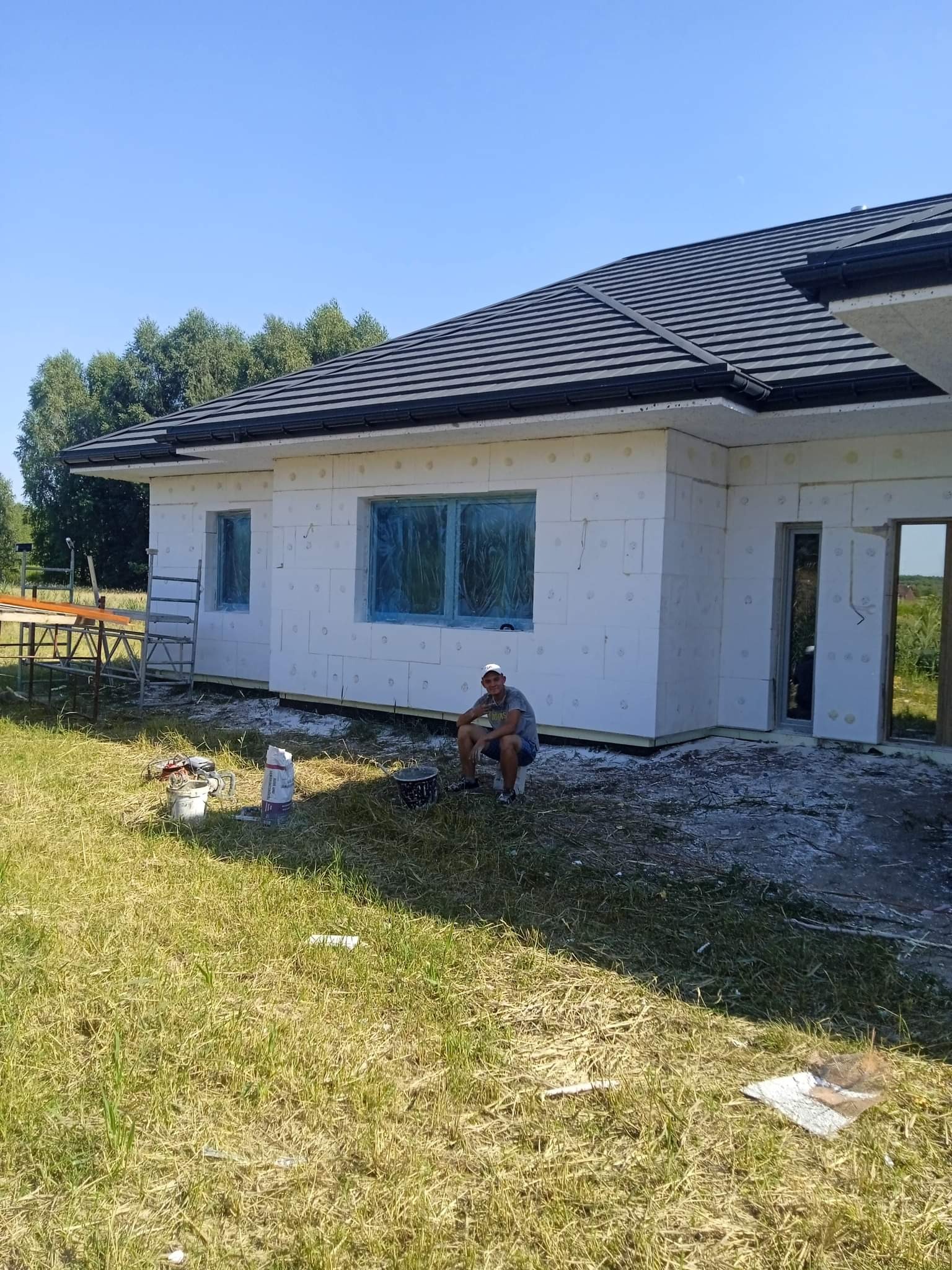 Dom jednorodzinny w trakcie prac: białe ściany z izolacją, zabezpieczone okna folią, mężczyzna siedzi na wiadrze przed budynkiem, obok leżą narzędzia i worek z materiałem.
