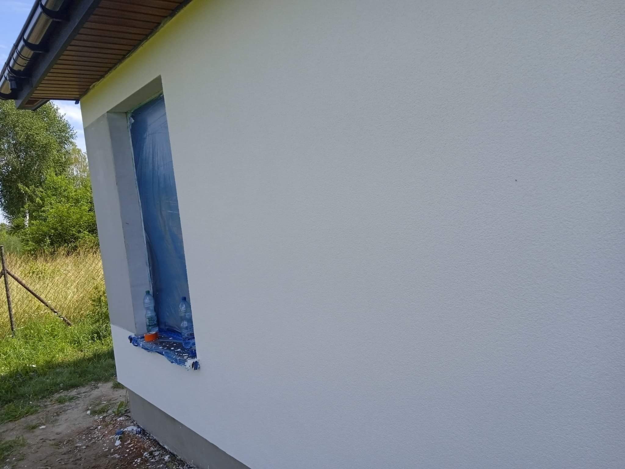 Fragment ściany budynku z oknem zabezpieczonym folią malarską, widoczne butelki z wodą na parapecie i ślady farby na ziemi, świeżo nałożony jasny tynk.
