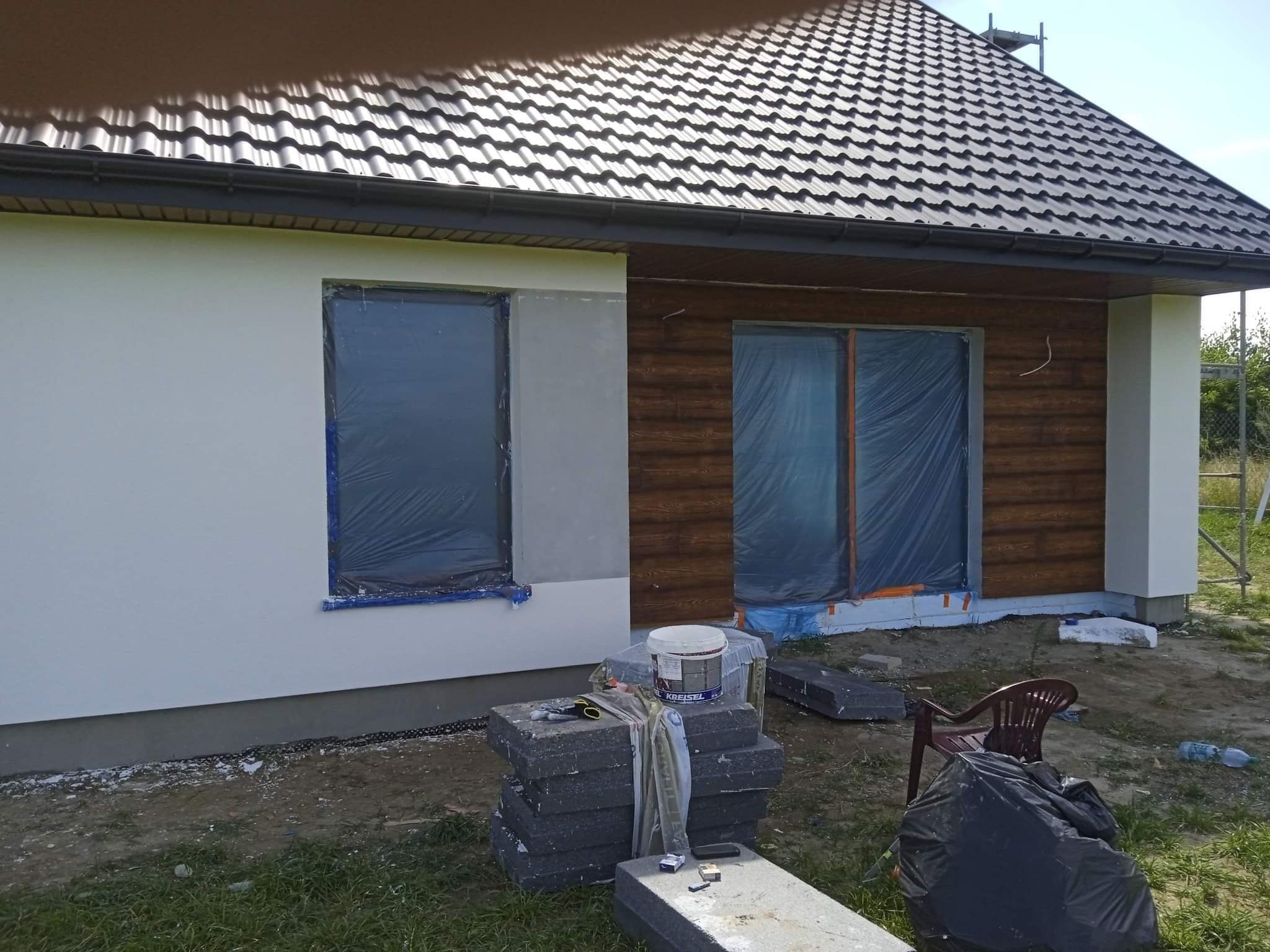 Dom jednorodzinny w trakcie ocieplania elewacji: fragment ściany otynkowanej, okna zabezpieczone folią, obok stos styropianu, wiadro z klejem Kreisel i narzędzia.