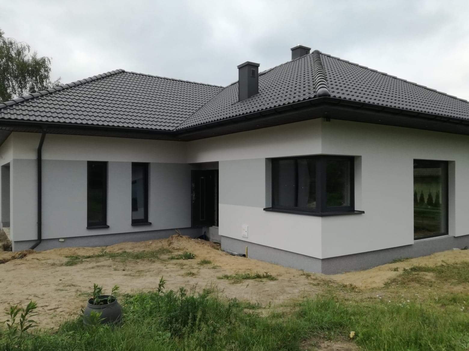 Nowoczesny dom jednorodzinny z szarą elewacją, ciemnymi oknami i grafitową dachówką, częściowo zagospodarowany teren wokół budynku.