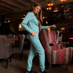 Kobieta prezentuje turkusowy dres z długim rękawem i dopasowanymi spodniami, stojąc w eleganckim wnętrzu restauracji z ręką w kieszeni i opierając się o krzesło w kolorowe paski.